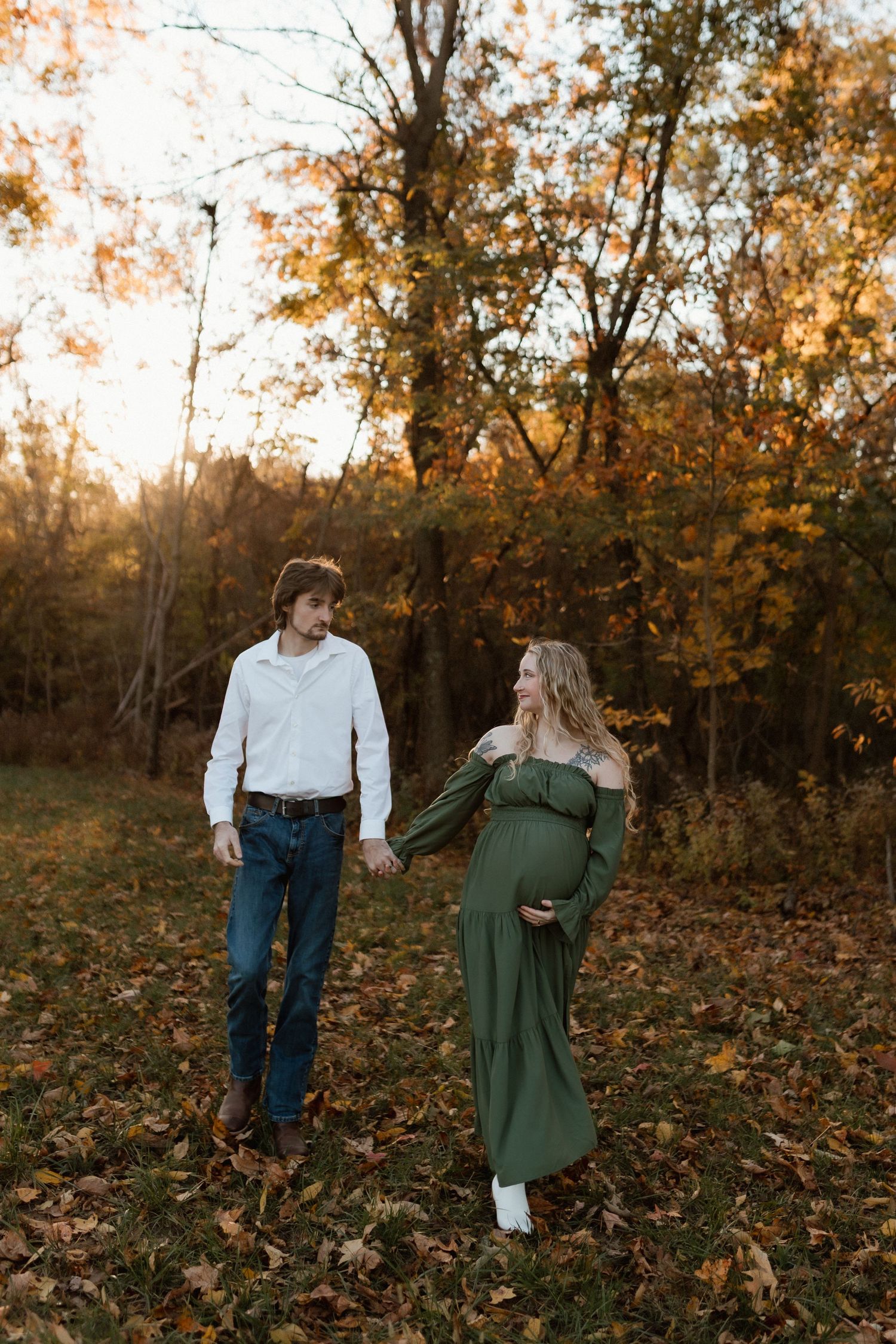 Couple holding hands in autumn forest, pregnant woman in green dress glows at sunset.