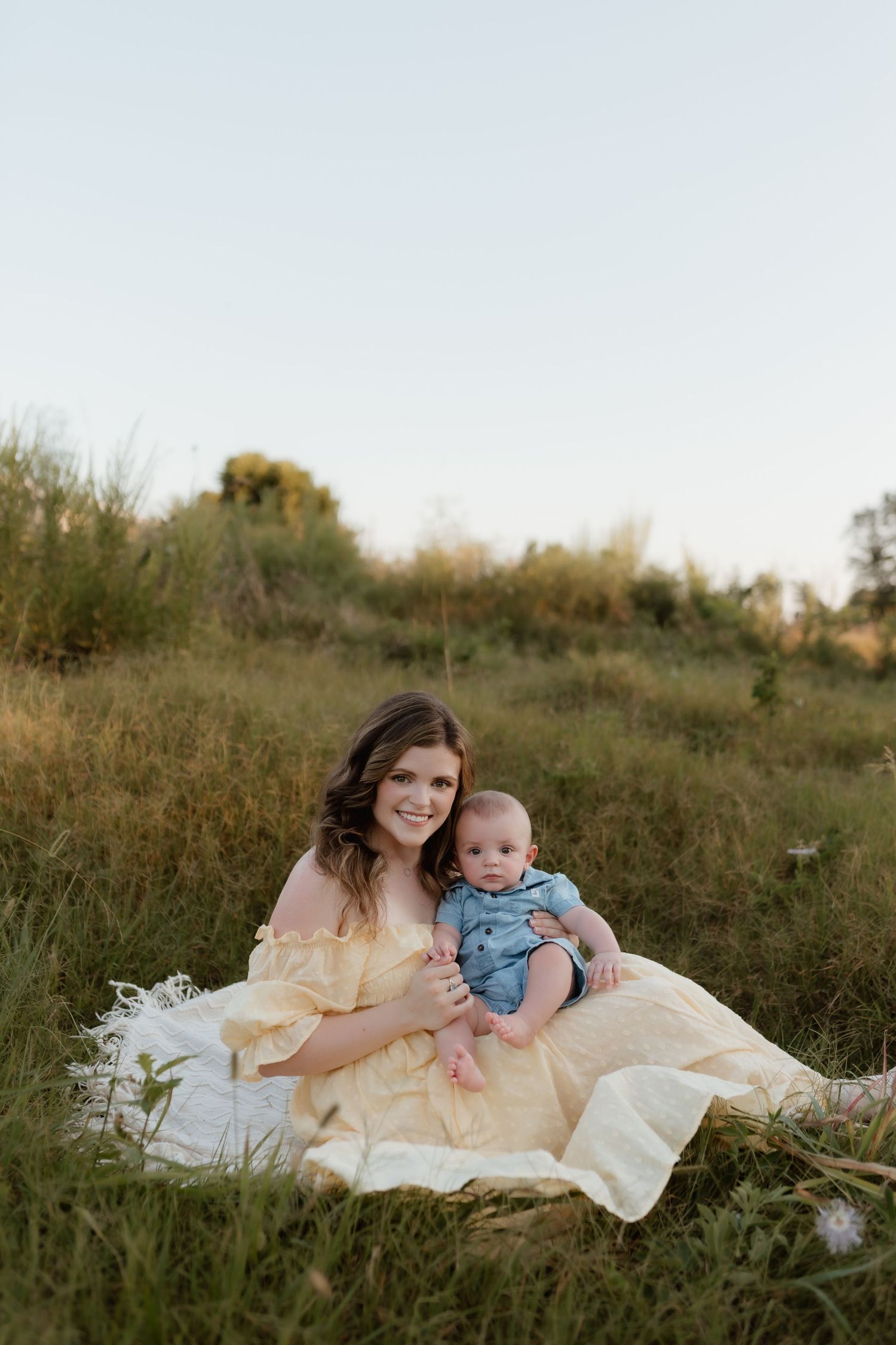 Smiling mother in yellow off-shoulder dress sitting on blanket in field with baby boy at sunset.