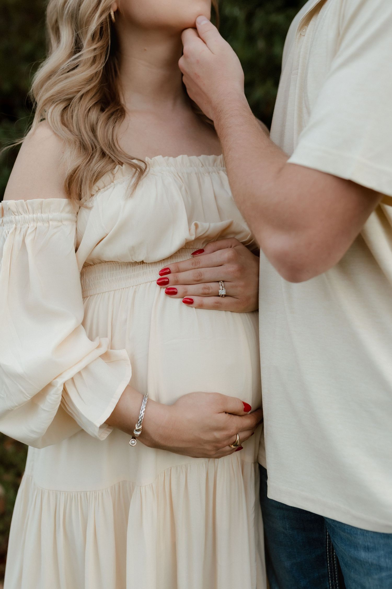 Close-up of pregnant couple holding baby bump, woman with red nails wearing cream dress.