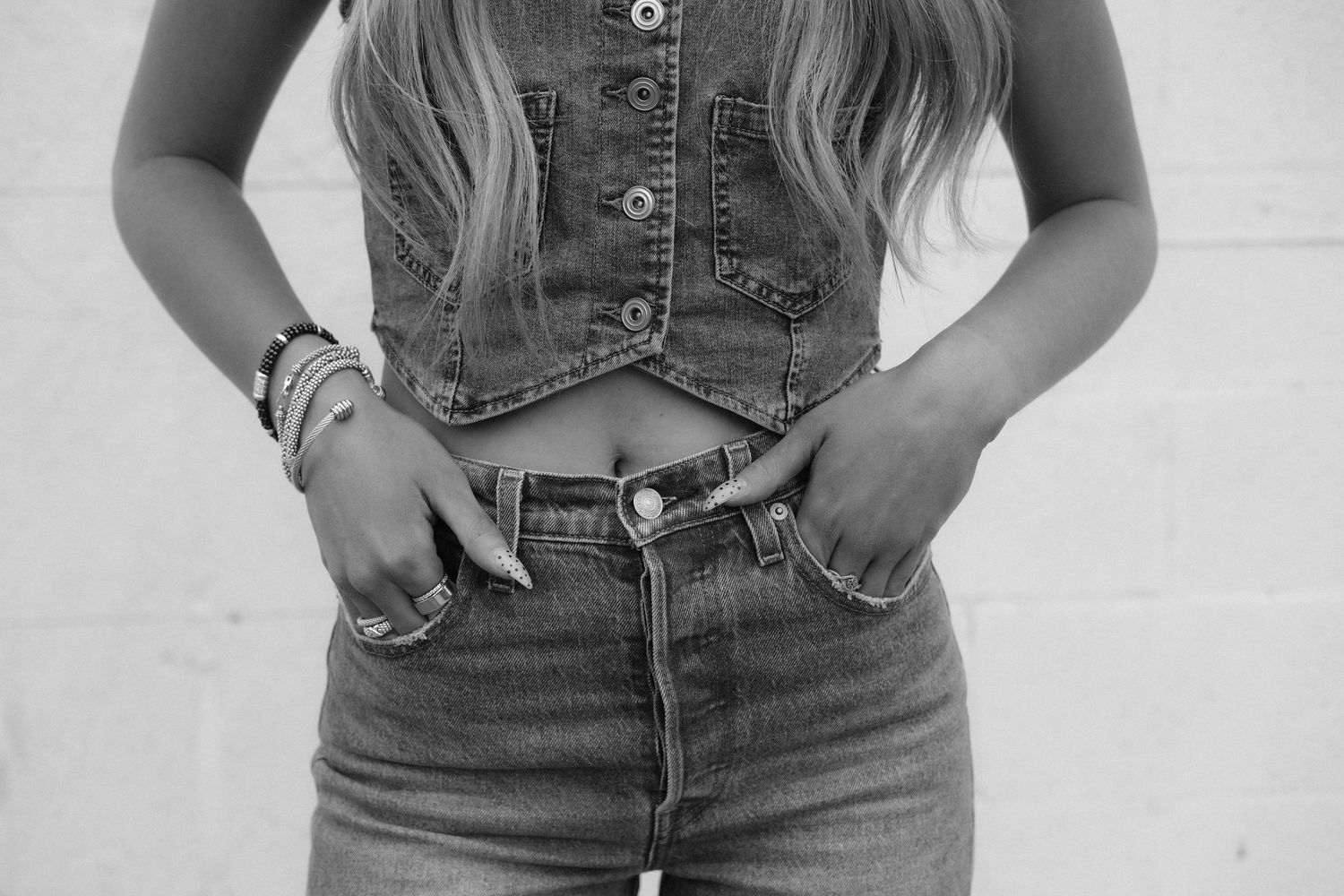 Black and white photo of a woman in a denim vest and high-waist jeans with bracelets and rings.