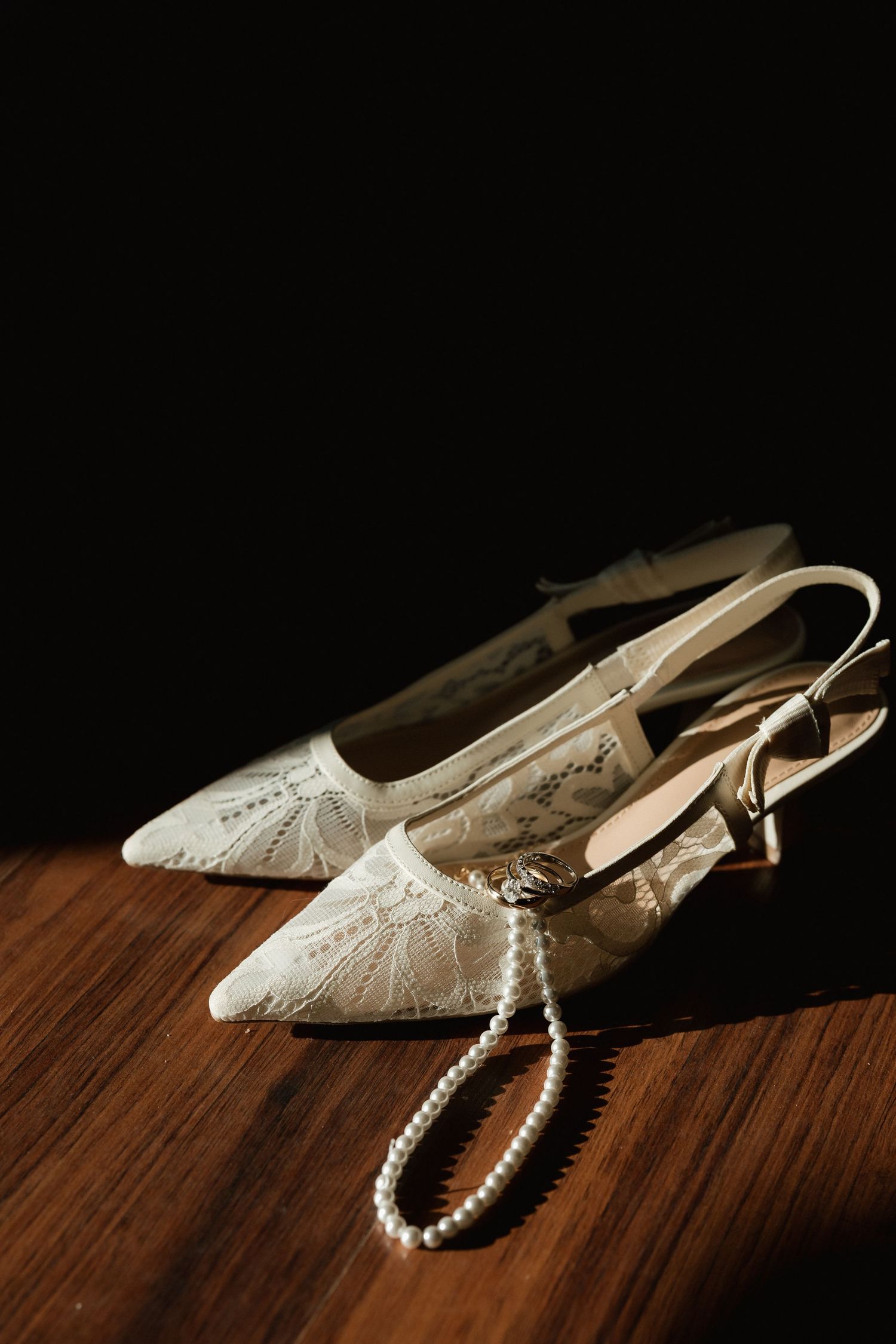 Ivory lace slingback bridal heels with pearl necklace on wooden surface against dark background.