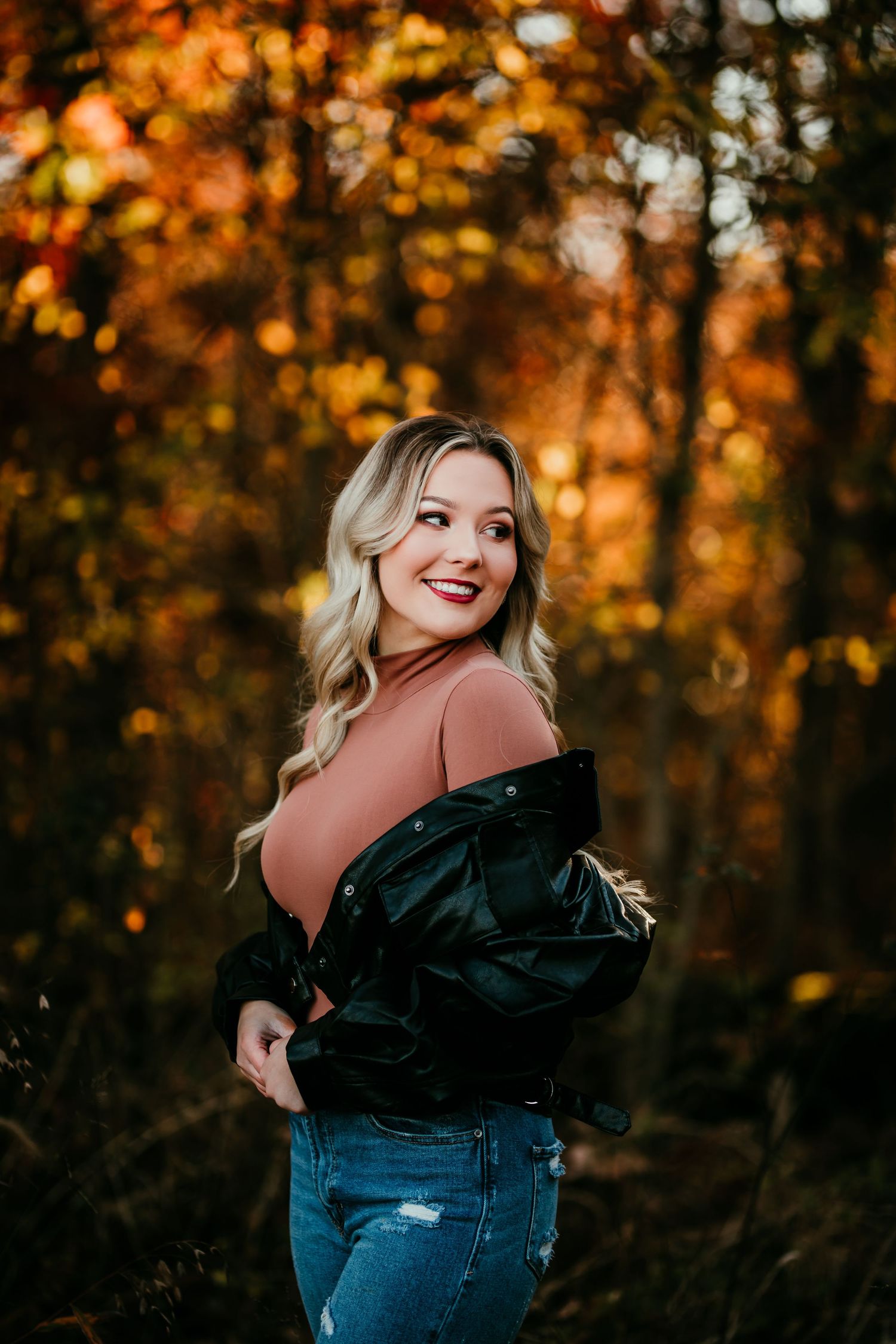 Blonde woman in black jacket and jeans posing in fall foliage.