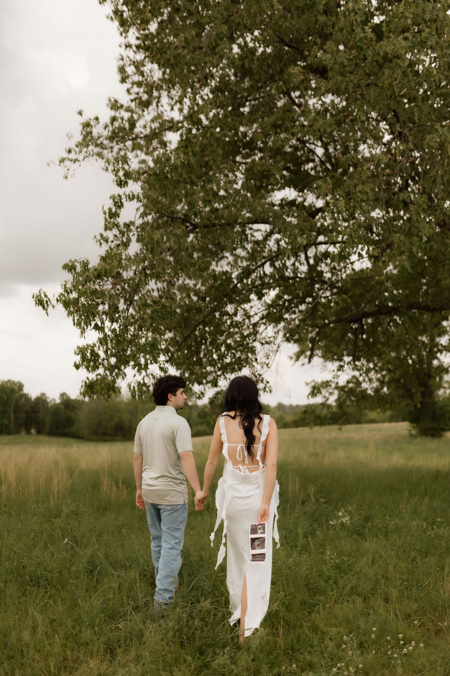 Pregnant couple walking hand in hand toward large tree in green field, woman in white lace dress holding ultrasound.