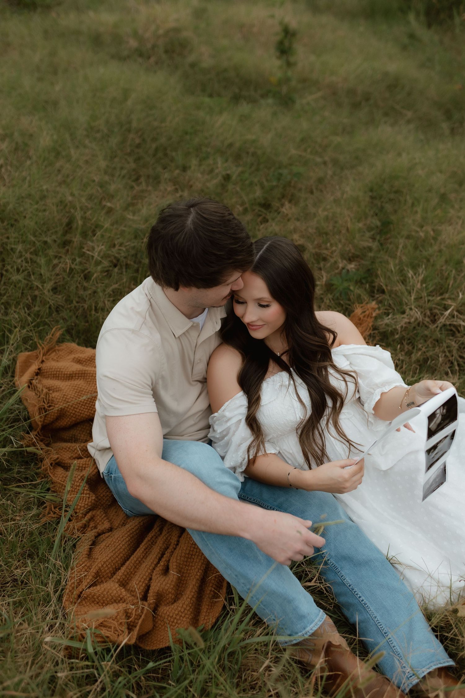 Couple sits on blanket in field holding ultrasound photo, sharing a sweet tender moment.
