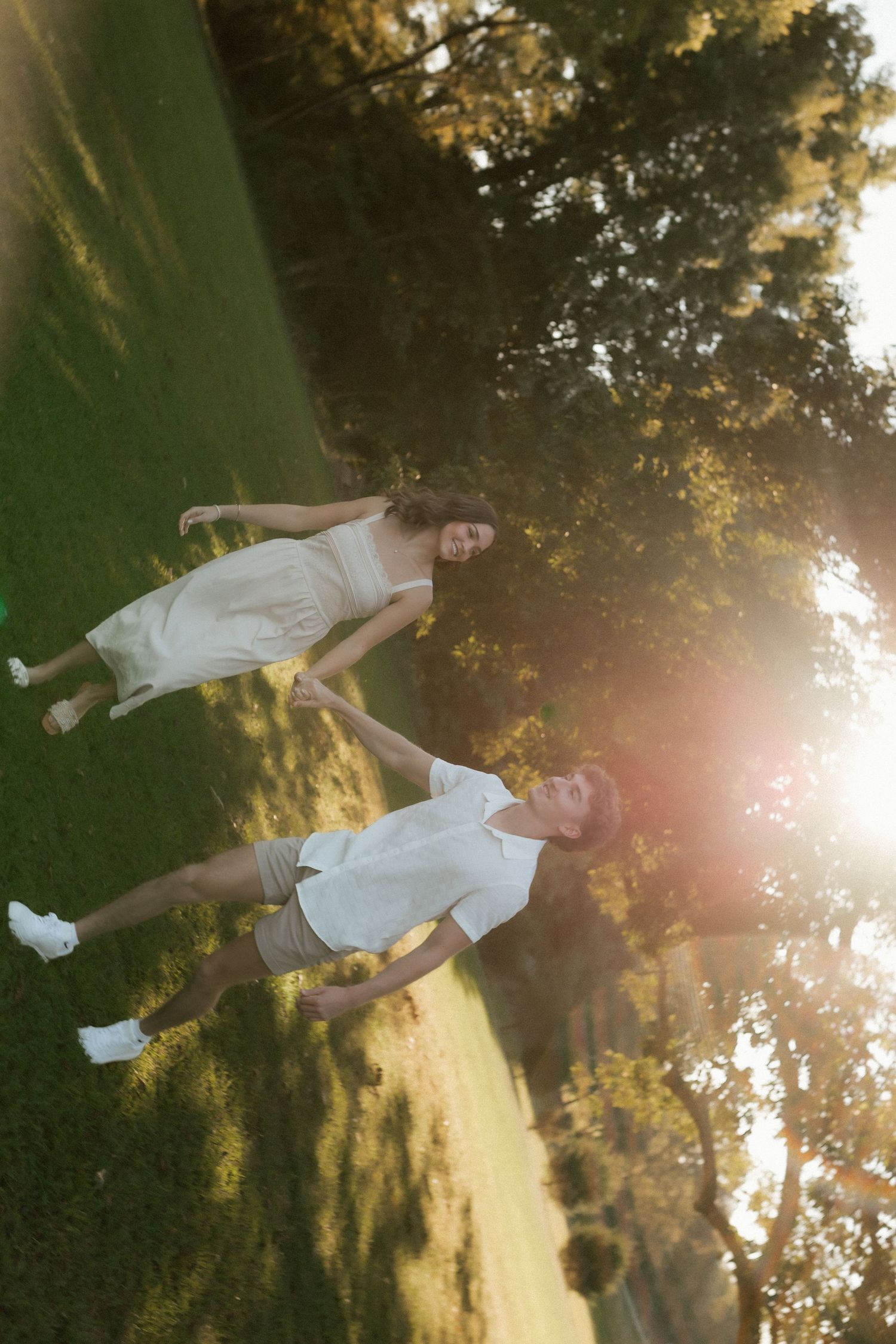 Aerial view of a couple walking hand in hand through a sunlit green park, both dressed in white casual outfits.