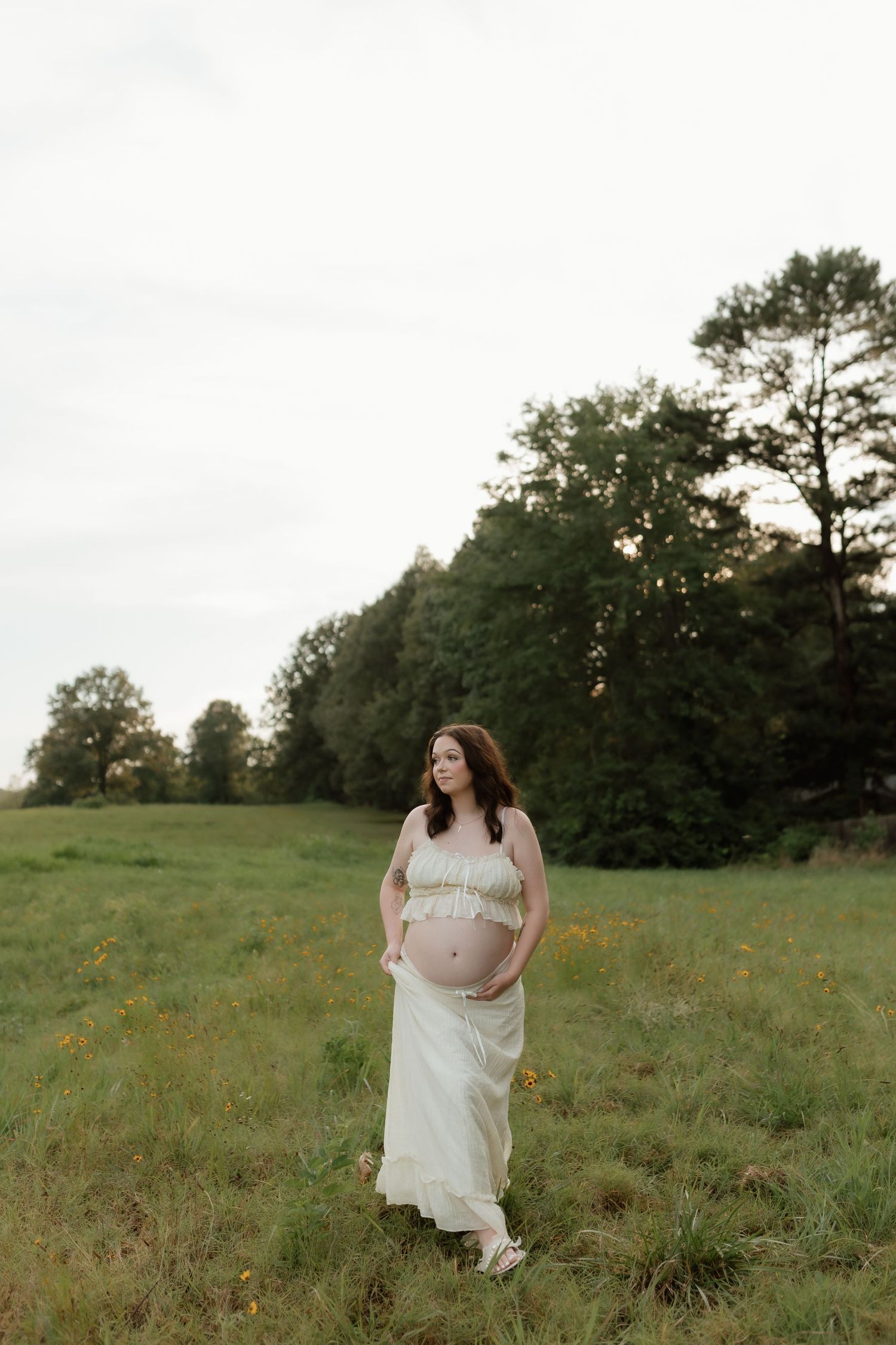 Pregnant woman in white two-piece maternity outfit standing in open green field at dusk.