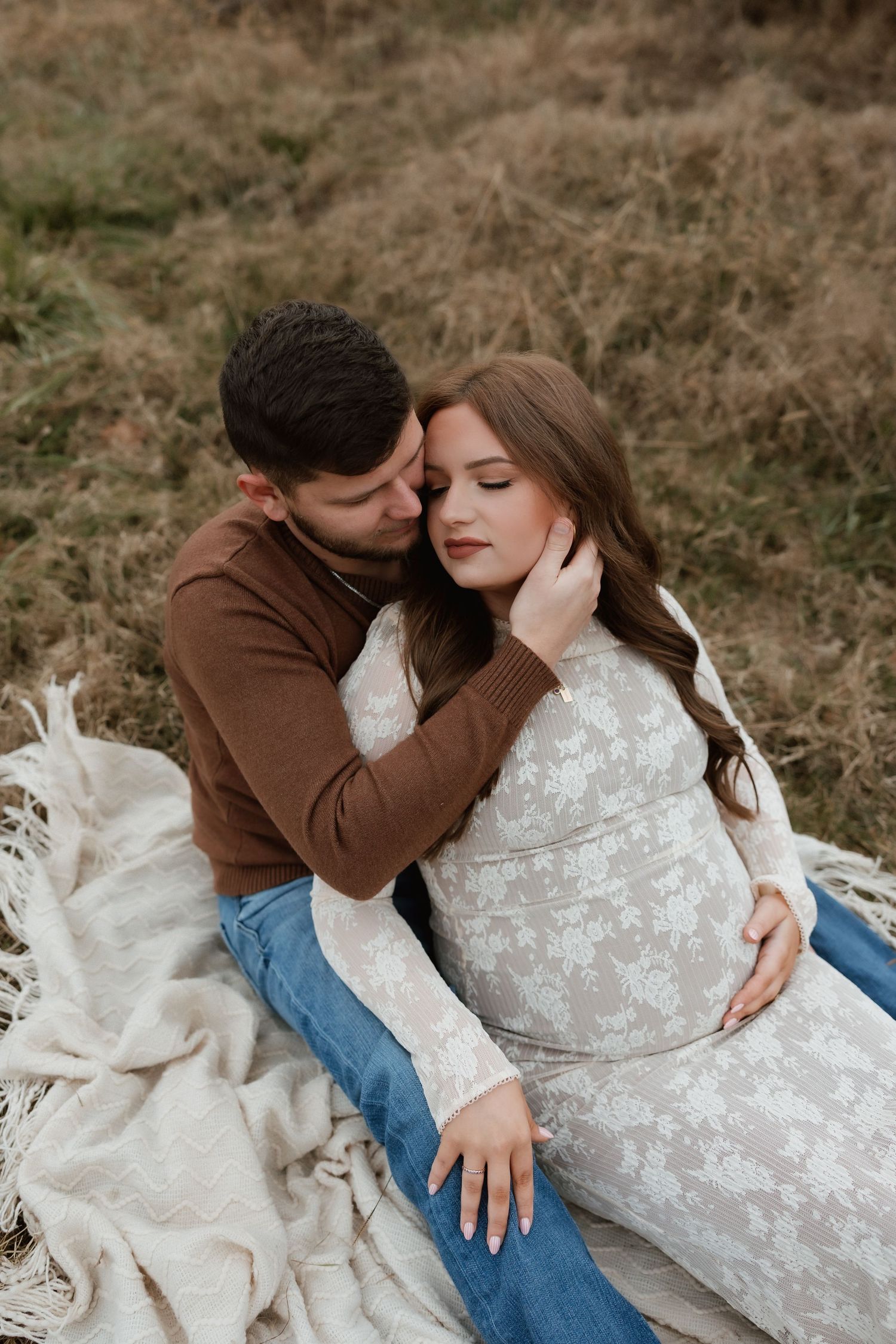 Couple cuddling on blanket in autumn field, man in brown sweater embracing pregnant woman in lace dress.