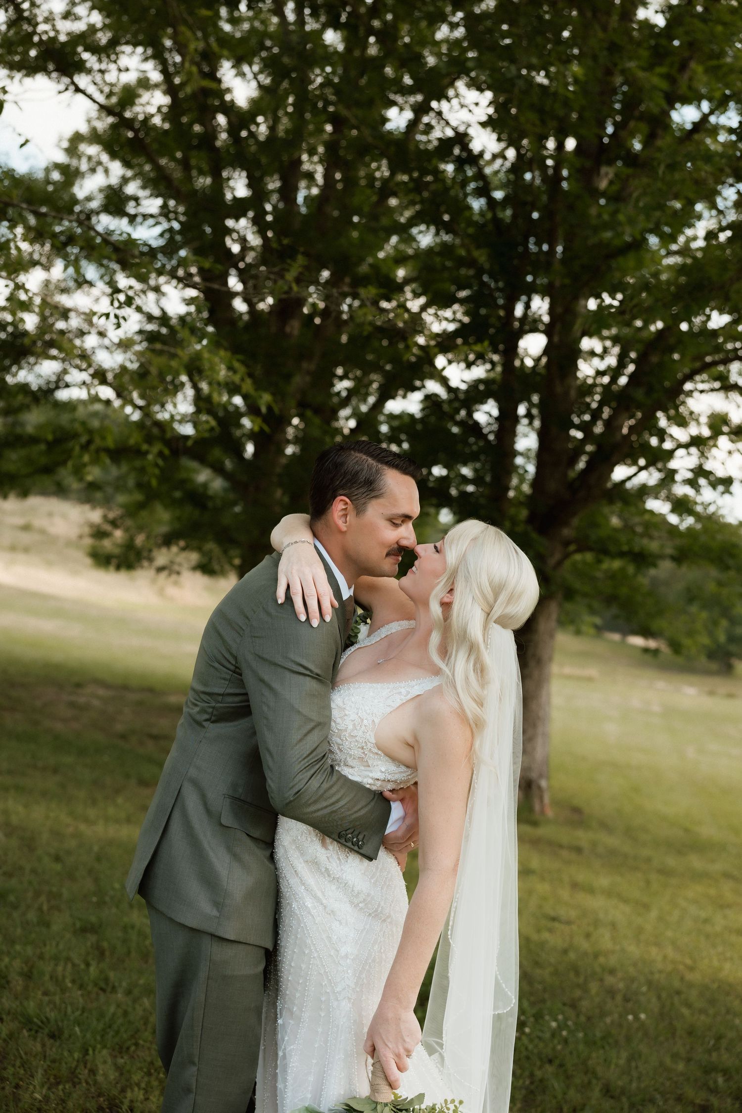 Bride in lace gown and groom in olive suit embracing near large tree in green meadow.