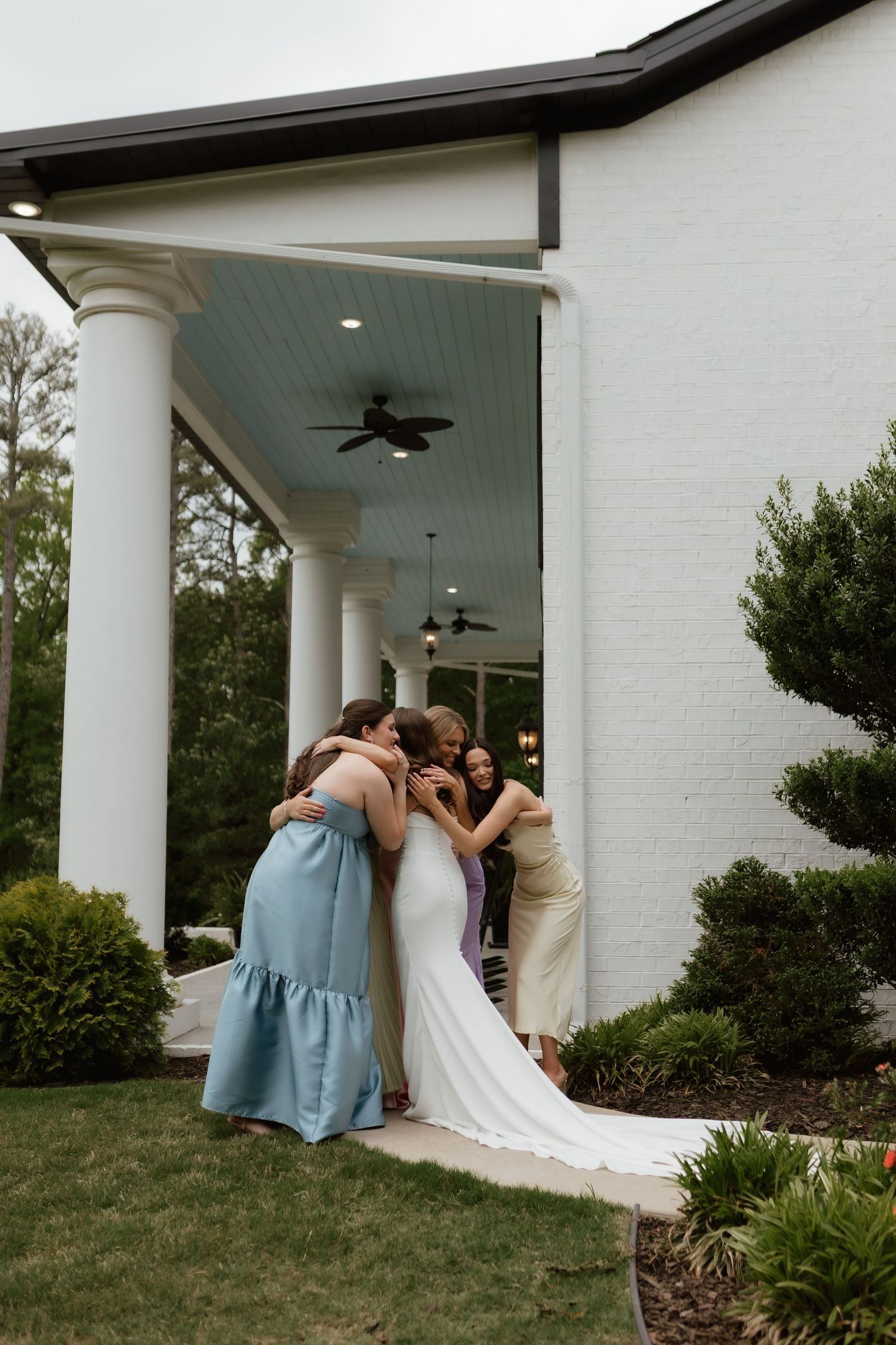Bride in white gown shares a group hug with bridesmaids outside a white colonial venue.