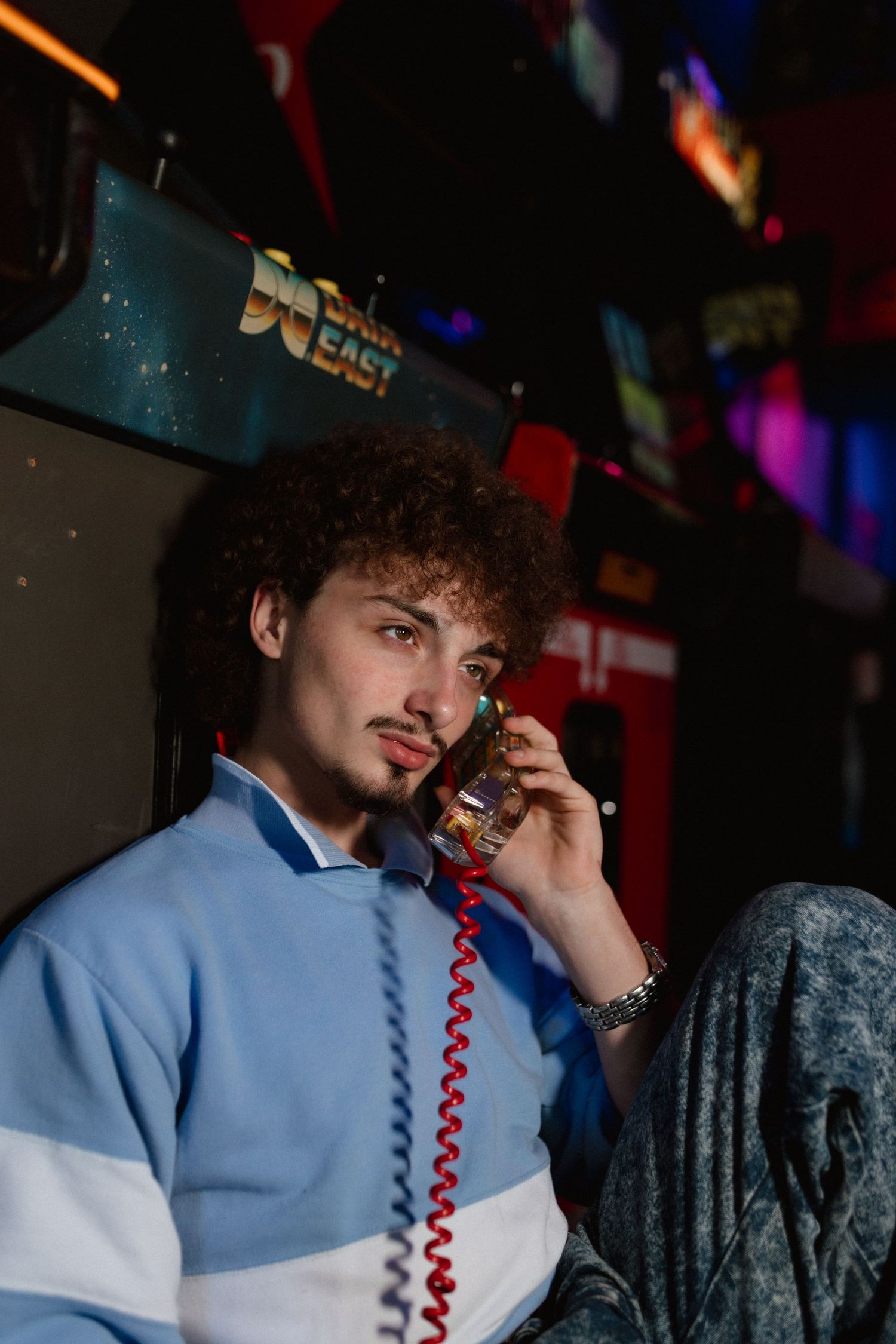 A young man with curly hair talks on a retro corded phone while sitting in a colorful arcade.