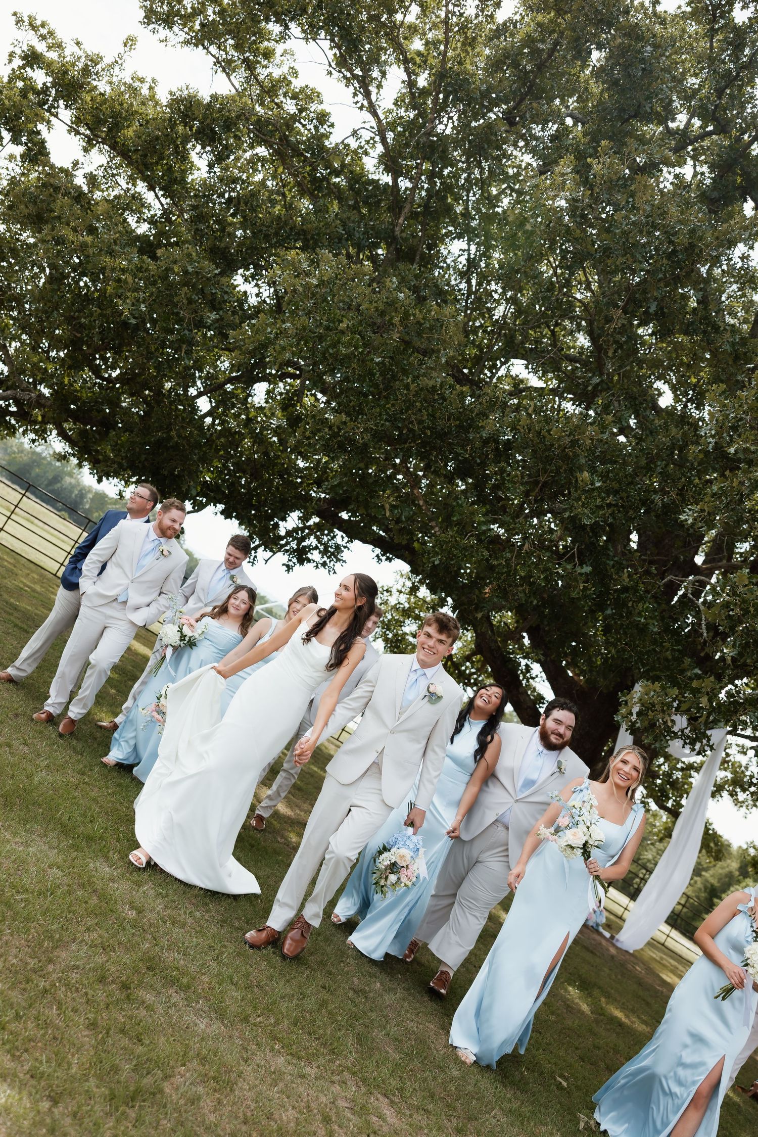 Wedding party in blue and beige attire walking together under large oak tree on lawn.