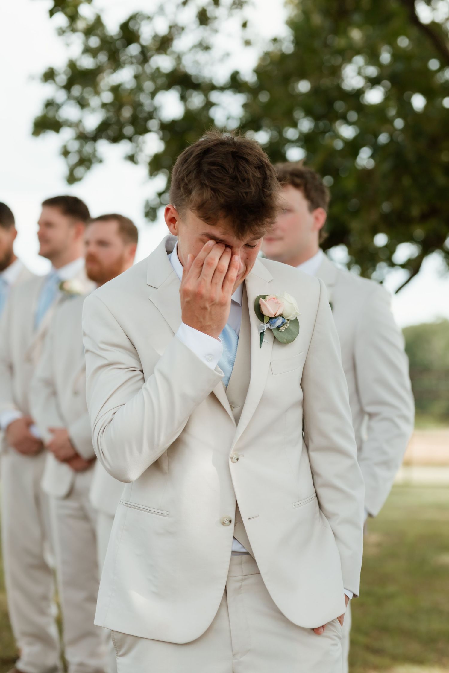 Emotional groom in beige suit wiping tears as bride walks down the aisle outdoors.