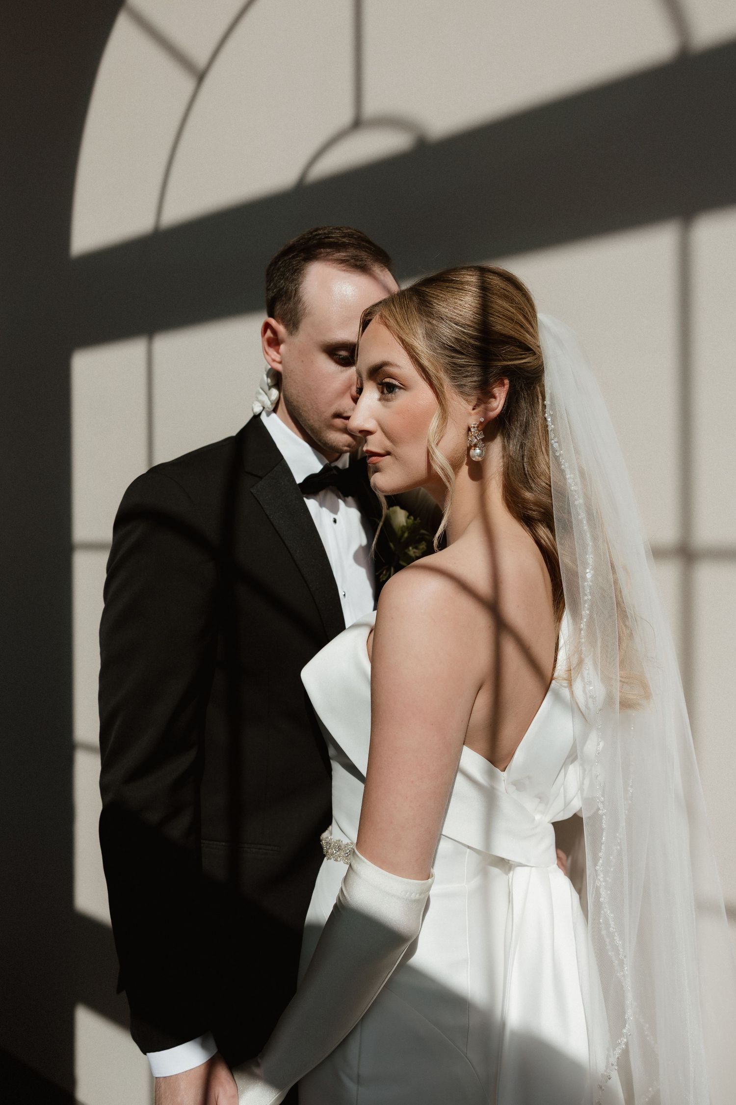 Bride and groom share an intimate moment in dramatic light and shadow indoors.