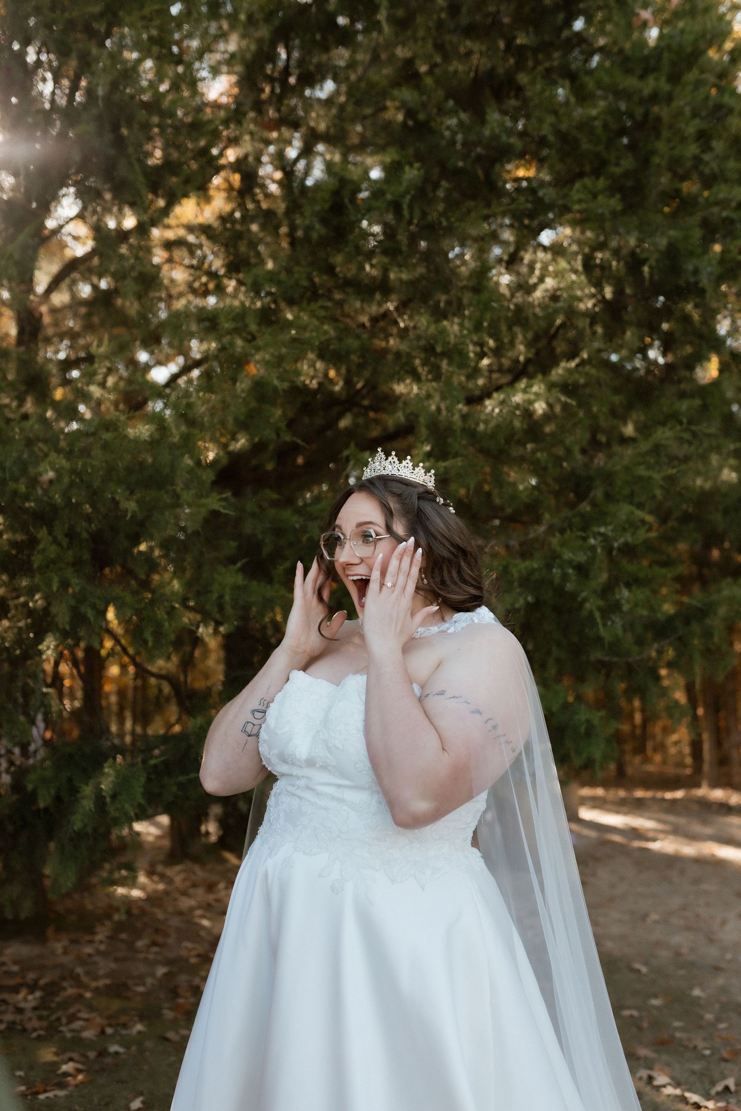 Bride in white gown and tiara poses with hands on face in a sunlit forest setting.