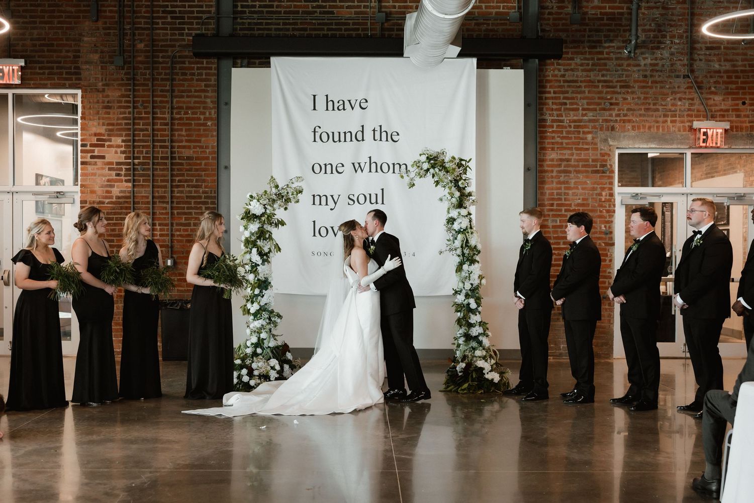 Bride and groom kiss at altar beneath romantic Song of Solomon quote wedding backdrop.