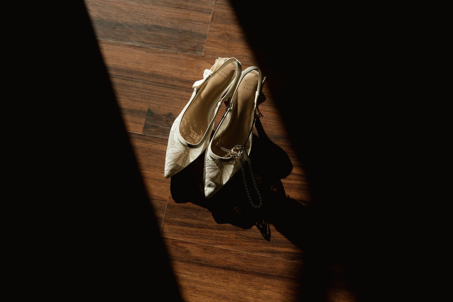 White bridal heels with ankle straps resting on wooden floor in dramatic shadow light.