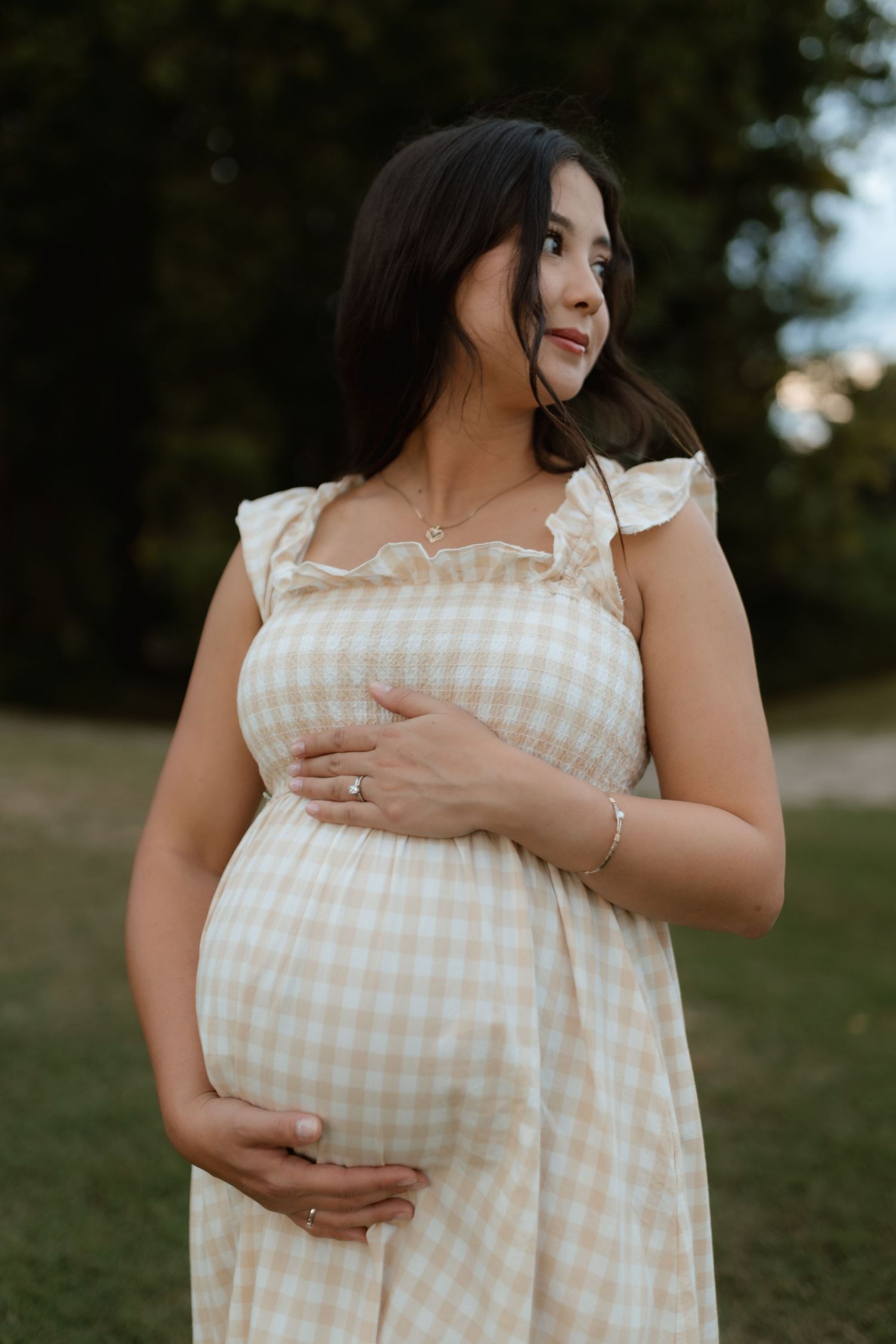 Pregnant woman in beige gingham dress cradling baby bump outdoors in a park setting.