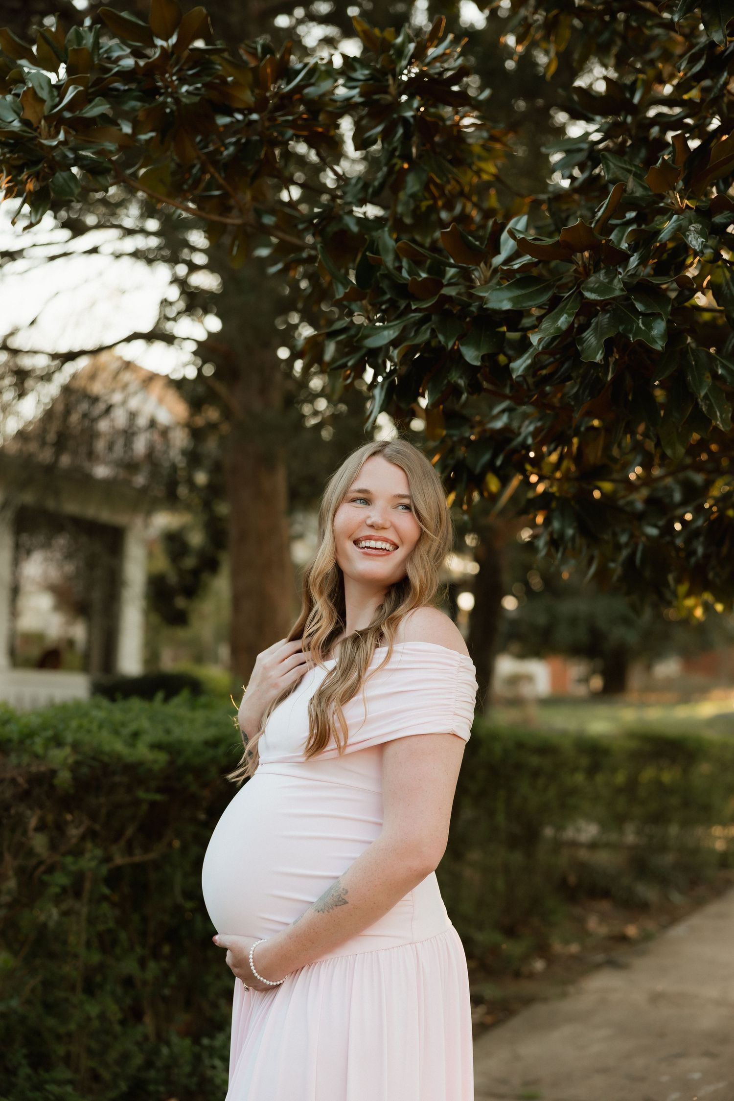 Smiling pregnant woman in a blush off-shoulder gown cradling her bump beneath a lush magnolia tree outdoors.