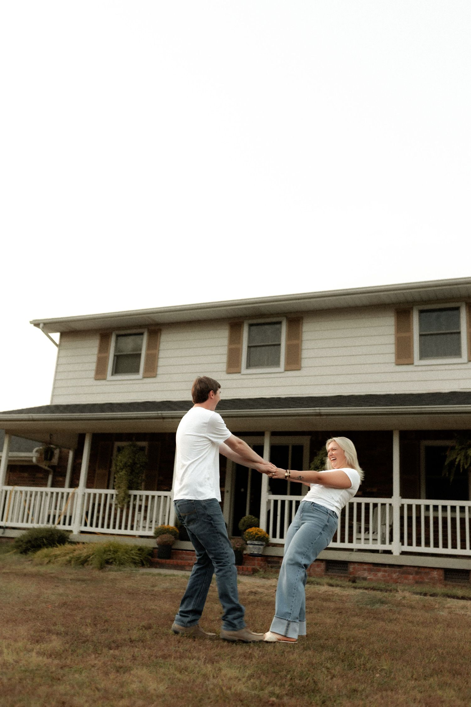 Happy couple dancing and holding hands in front yard of two-story white farmhouse with covered porch.