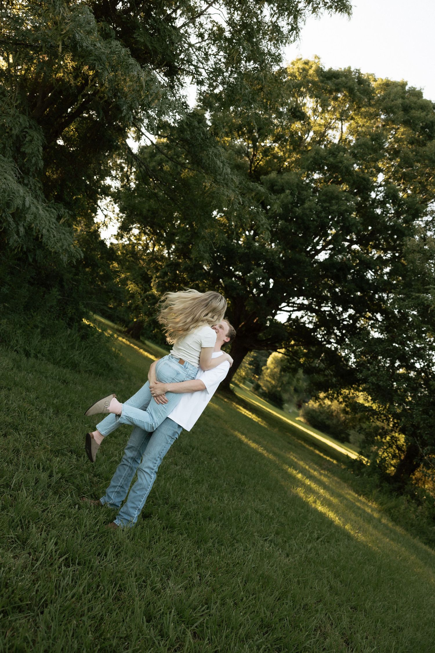 A romantic couple embracing and kissing on a sunlit grassy hillside beneath a large tree at sunset.