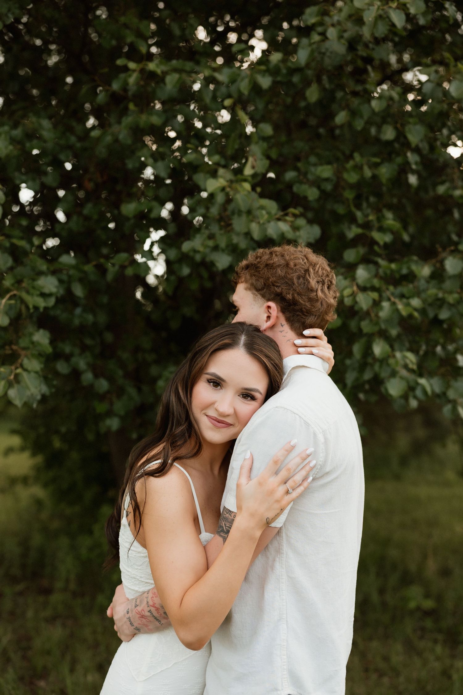 A smiling woman with tattoos embraces a curly-haired man near a lush green tree outdoors.