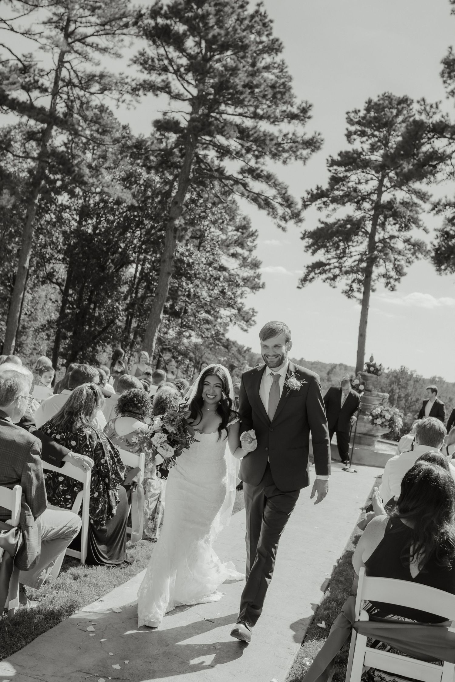 Black and white photo of a bride and groom walking down the aisle at an outdoor wedding ceremony surrounded by tall pine trees.