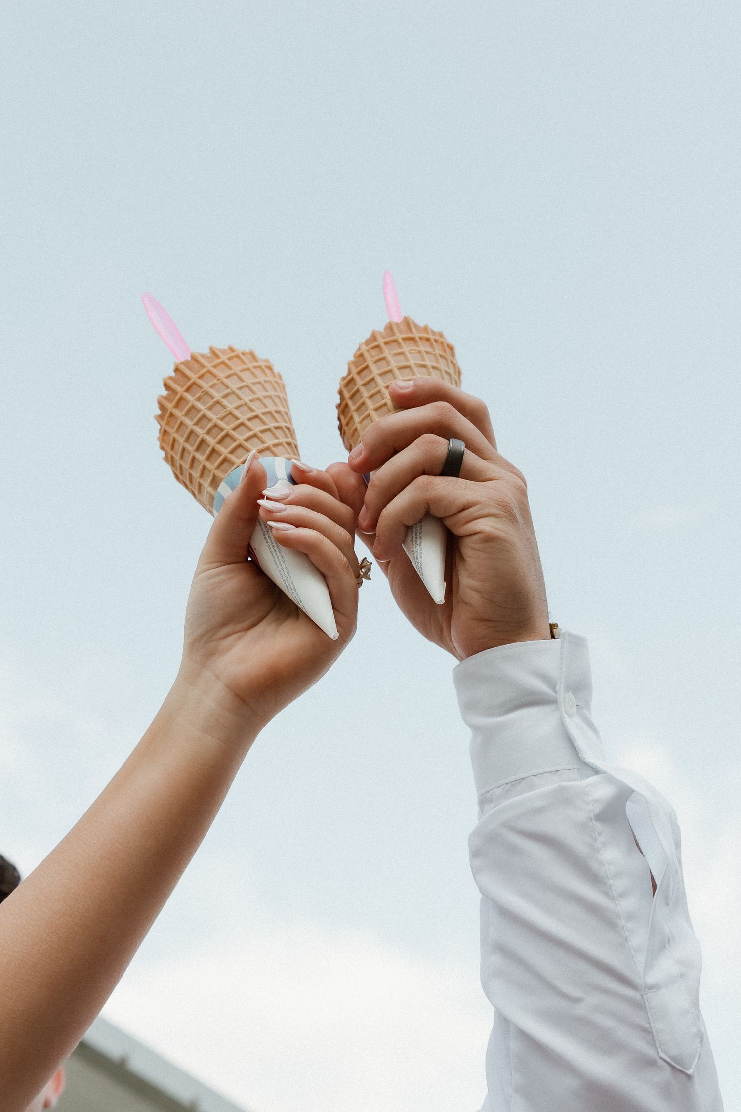 Two hands raise waffle ice cream cones together against a light sky in a celebratory gesture.