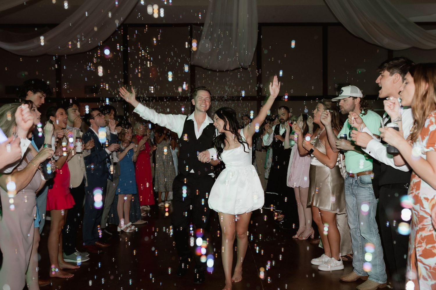 Bride and groom celebrate on the dance floor surrounded by bubbles and cheering wedding guests.