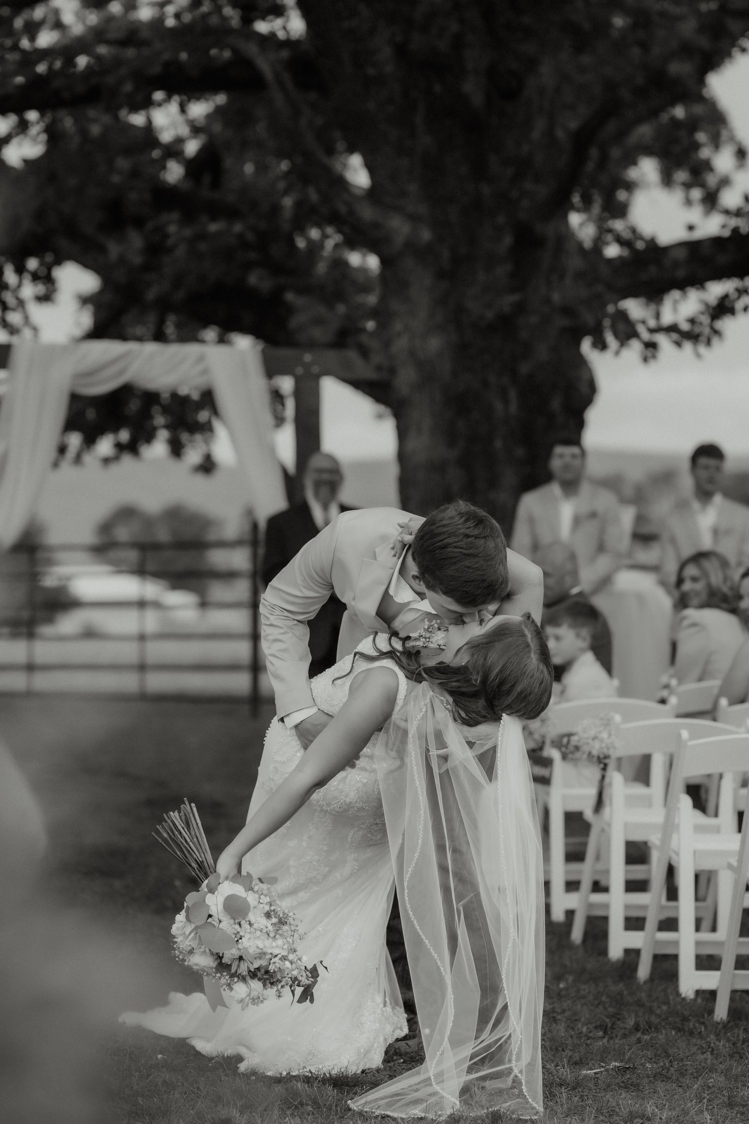 Black and white photo of groom dipping bride for a kiss at outdoor wedding ceremony under large tree.