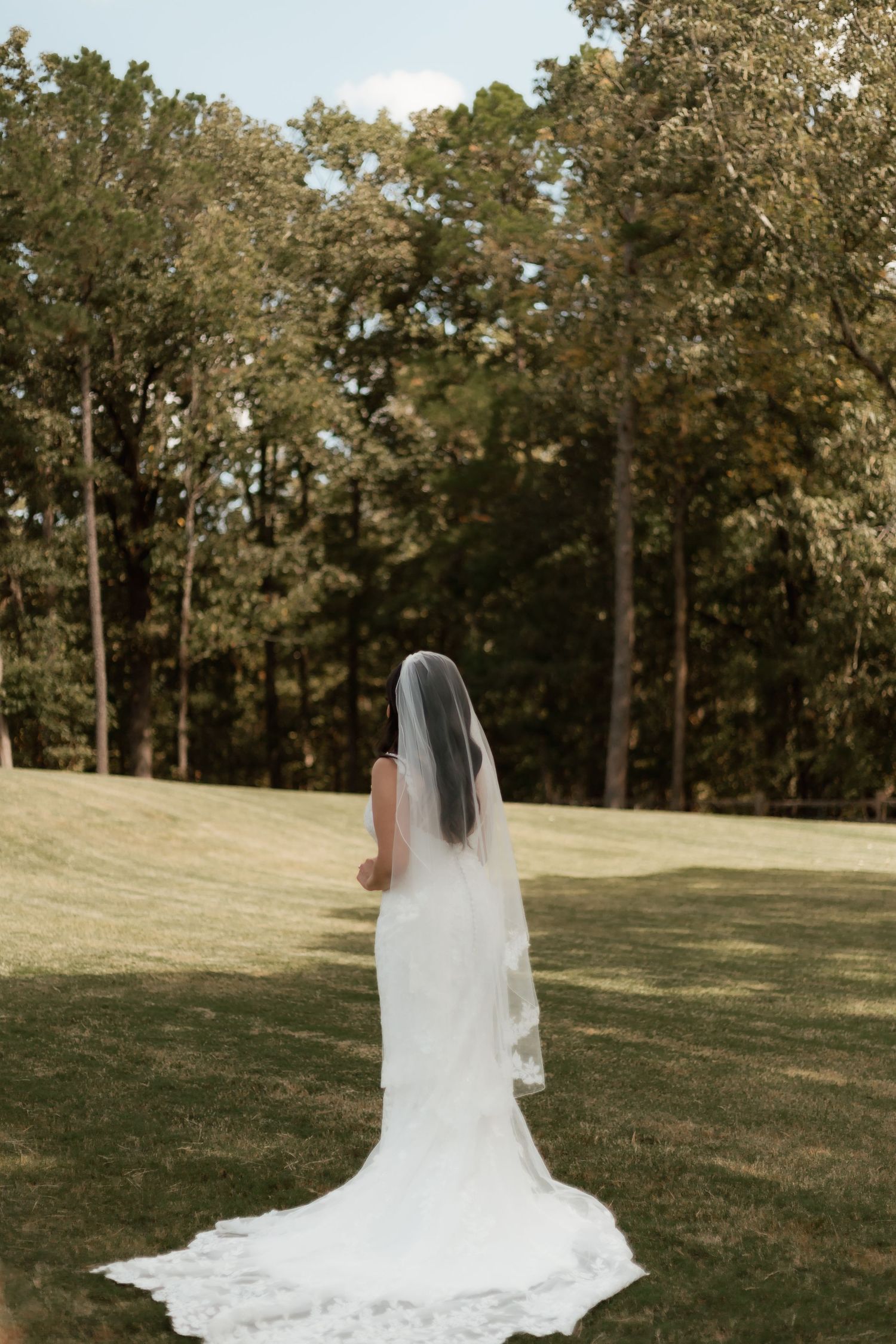 Bride in elegant white wedding gown with long veil standing on green lawn facing a forest.