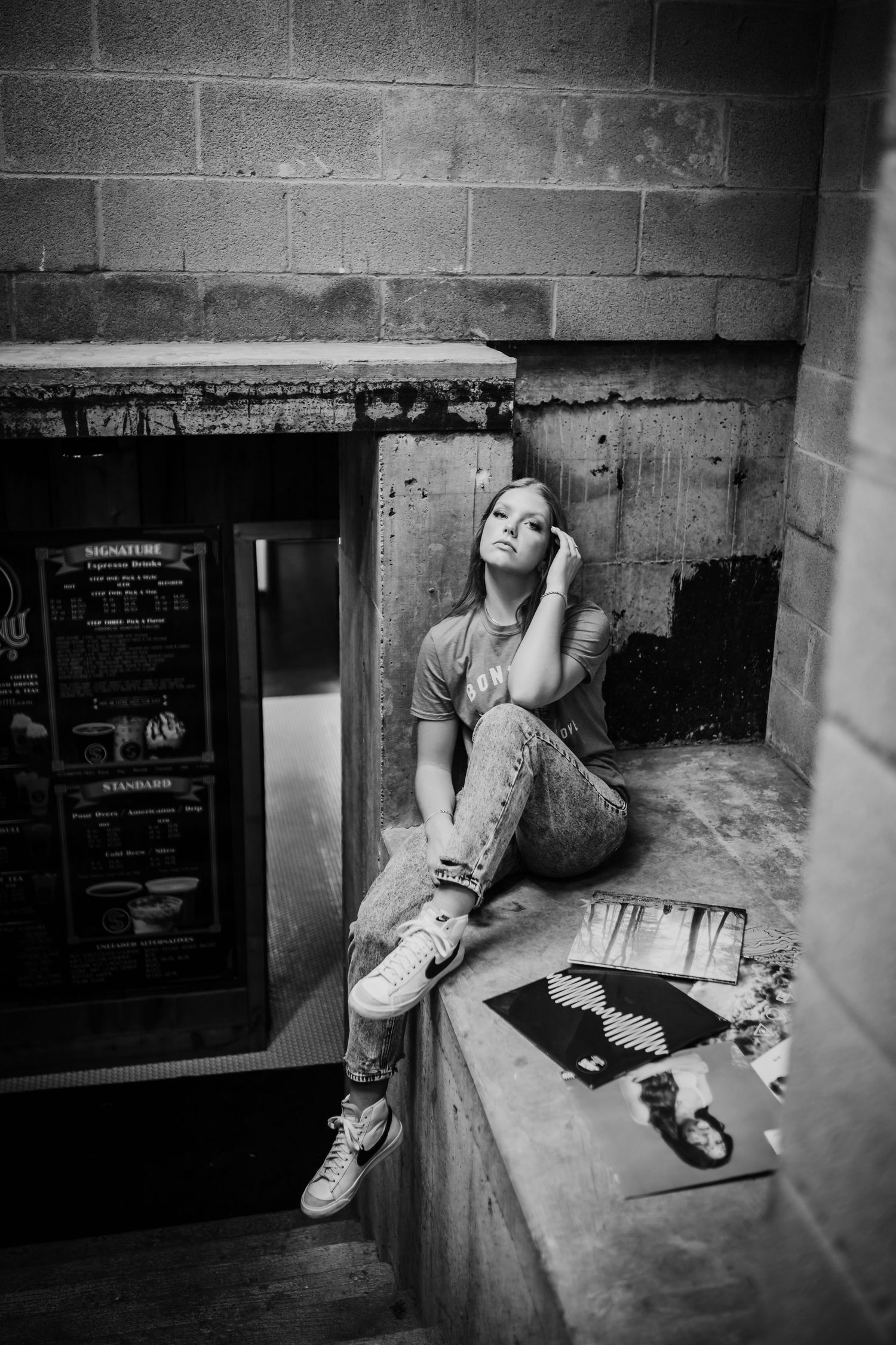 Black and white photo of teenage girl sitting on concrete ledge outside urban building.