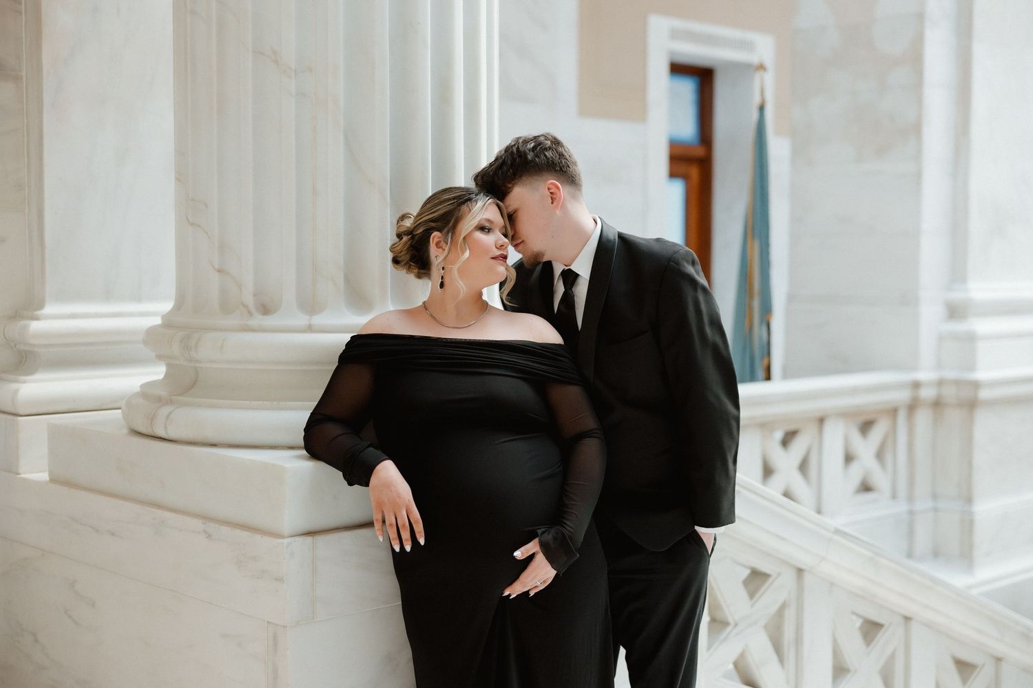 Couple sharing intimate moment, pregnant woman in black dress embraced by partner near marble columns.