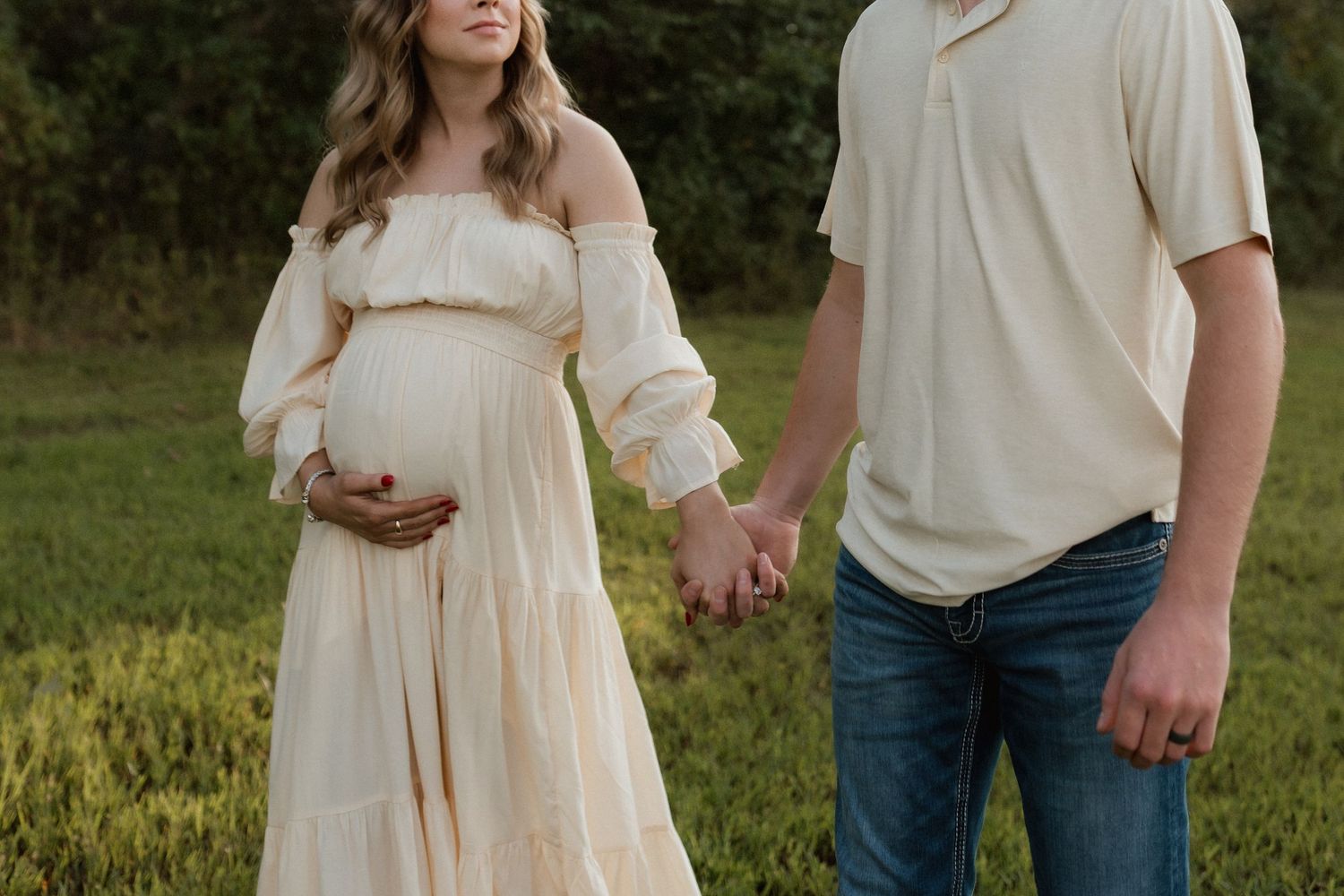 Pregnant woman in cream off-shoulder gown holds belly while partner holds her hand outdoors.