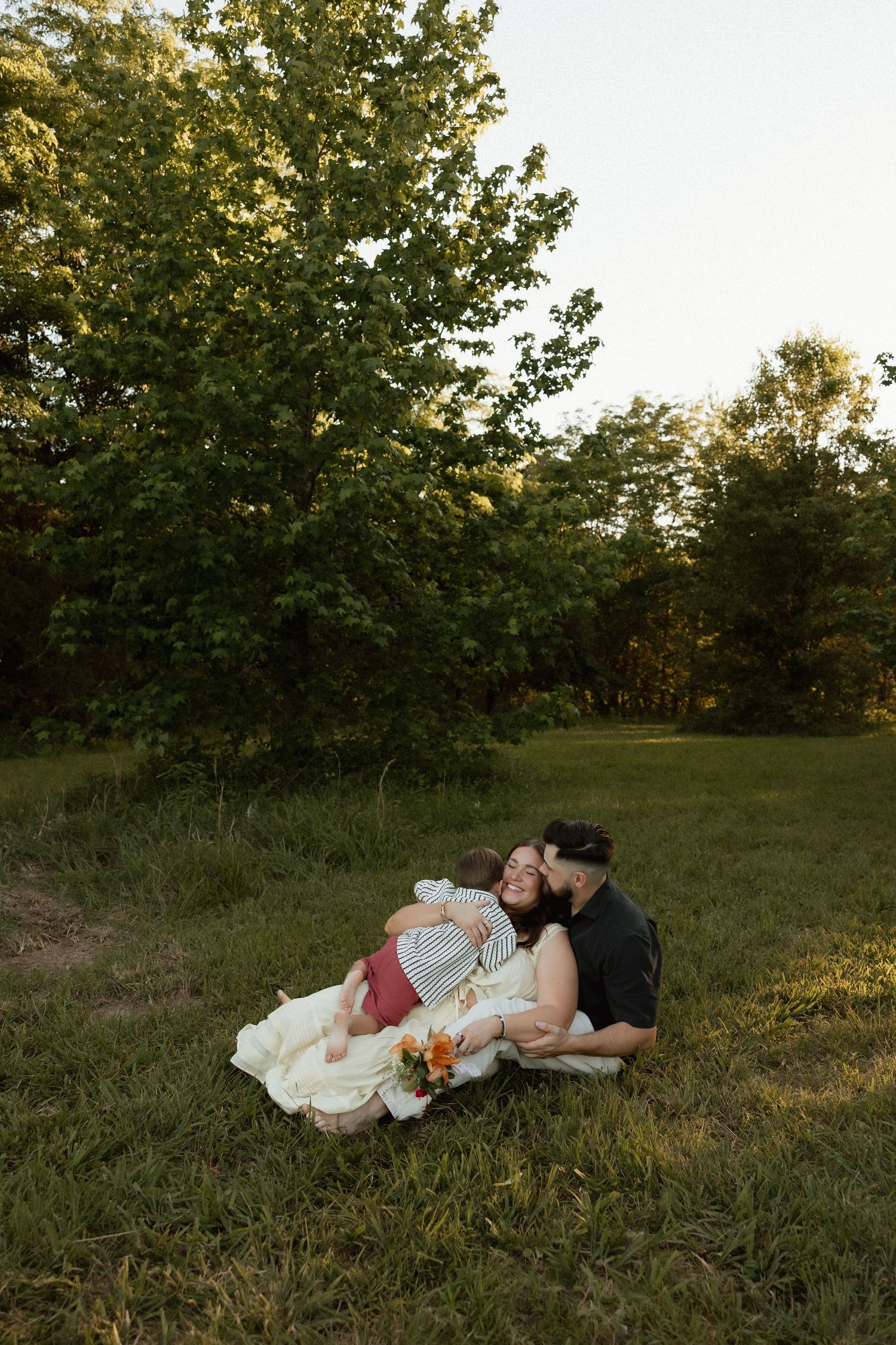 Family of three sitting together on green grass with man kissing woman while she holds baby.