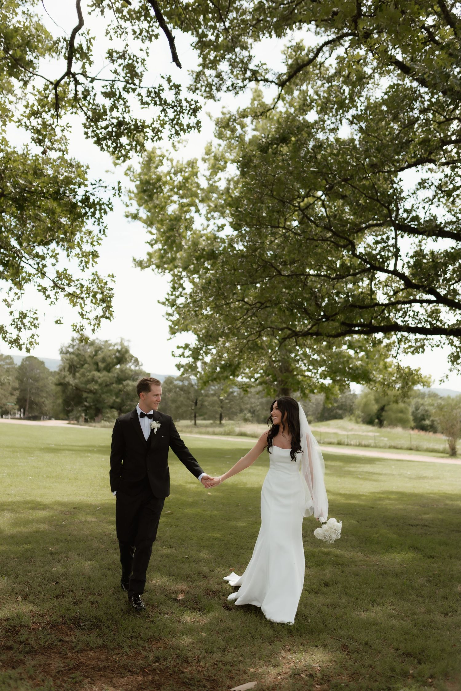 Bride and groom holding hands walking under large oak tree in lush green park setting.