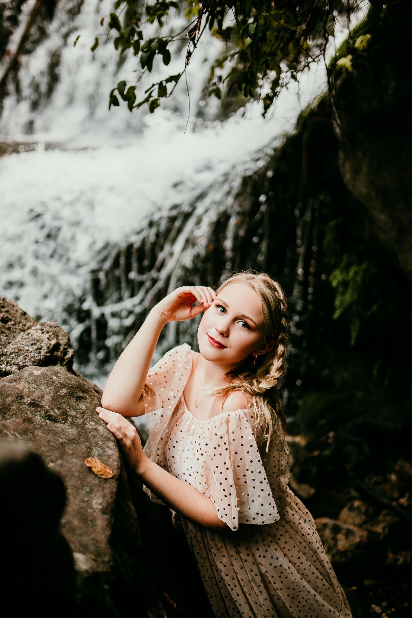 Blonde woman in polka dot dress poses on rocks near cascading waterfall in lush forest setting.