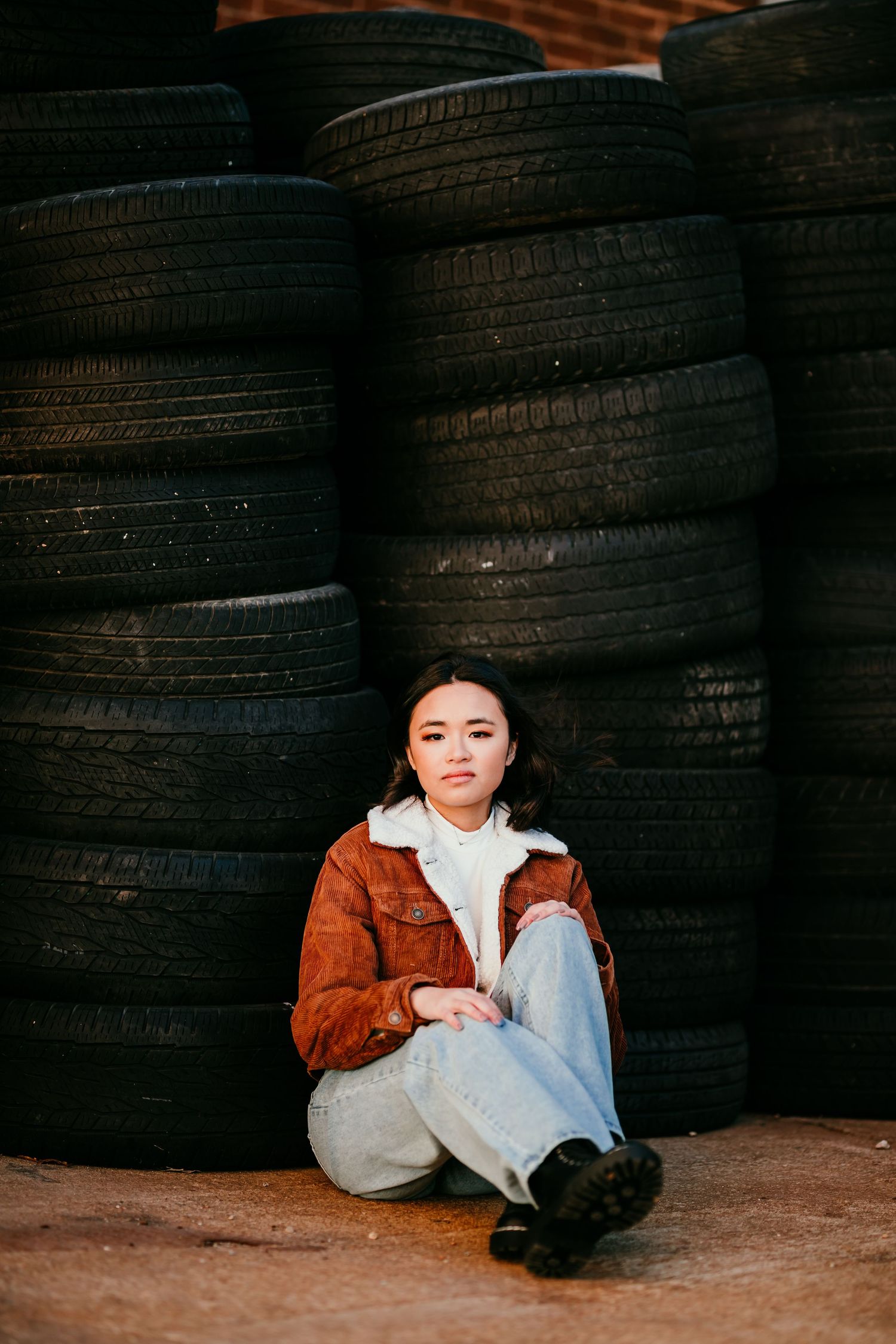 Young Asian woman in brown sherpa jacket sitting cross-legged in front of stacked black tires outdoors.