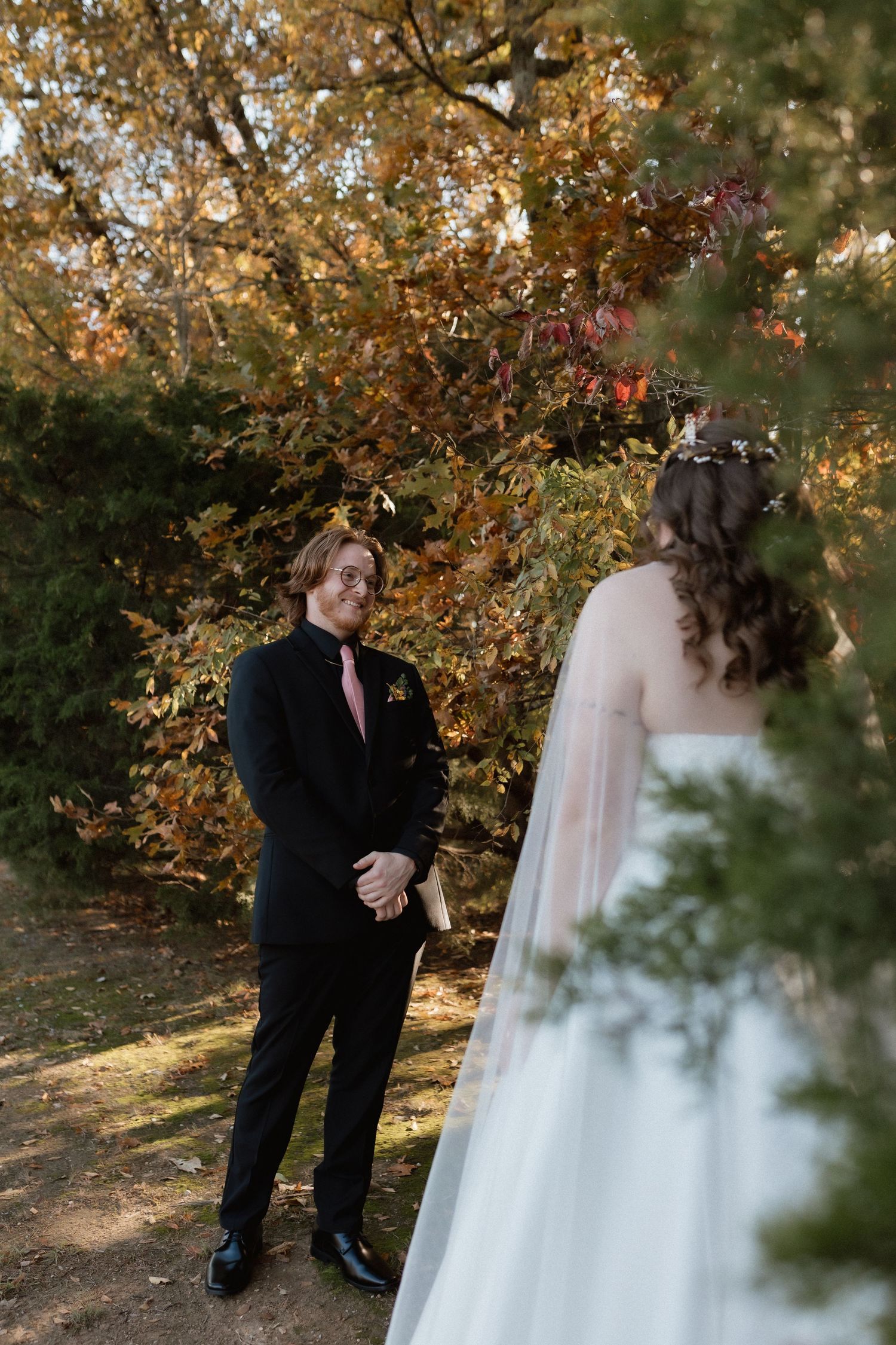 Groom in black suit smiles at bride during romantic first look moment among autumn foliage.