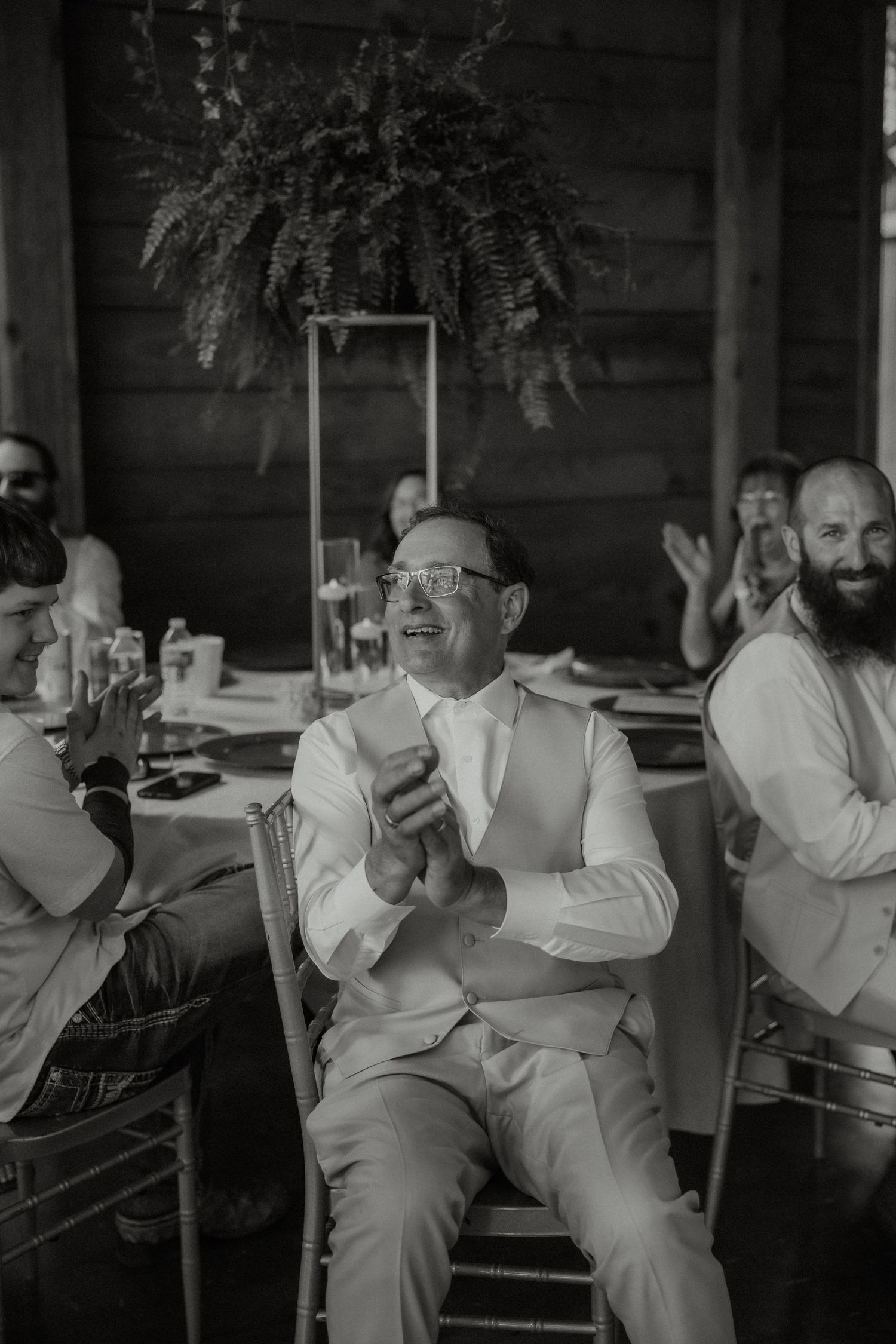 Groom in white suit clapping and laughing at wedding reception with guests in rustic barn.