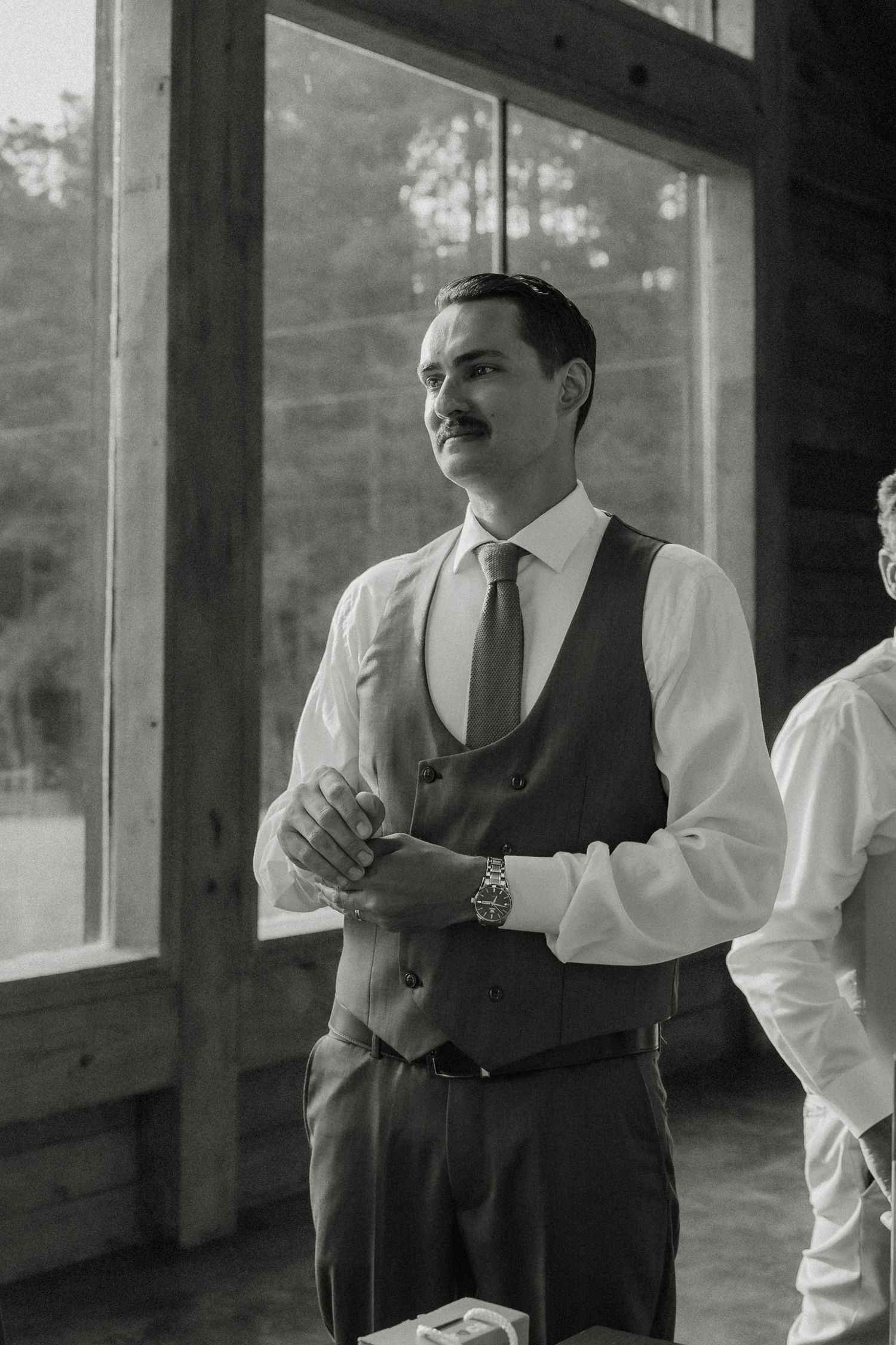 Black and white portrait of dapper groom in vest and tie adjusting cufflinks near large wooden framed windows.