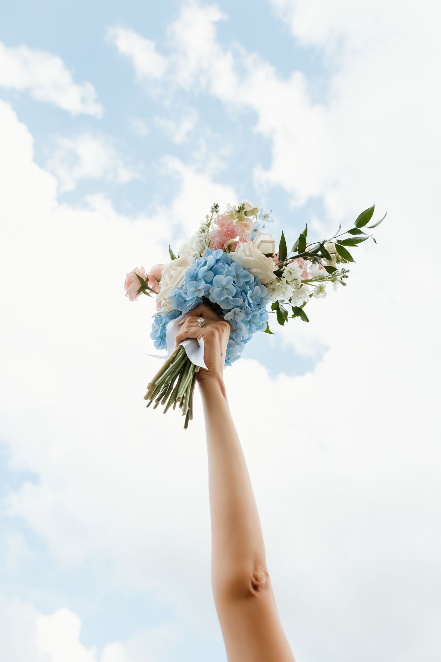 Bride holding blue hydrangea and pink floral wedding bouquet raised against bright cloudy sky.