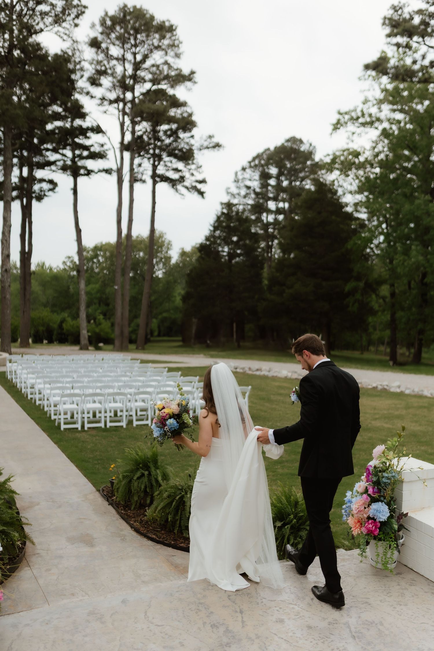 Groom escorts bride in flowing veil past rows of white chairs at outdoor wedding venue.