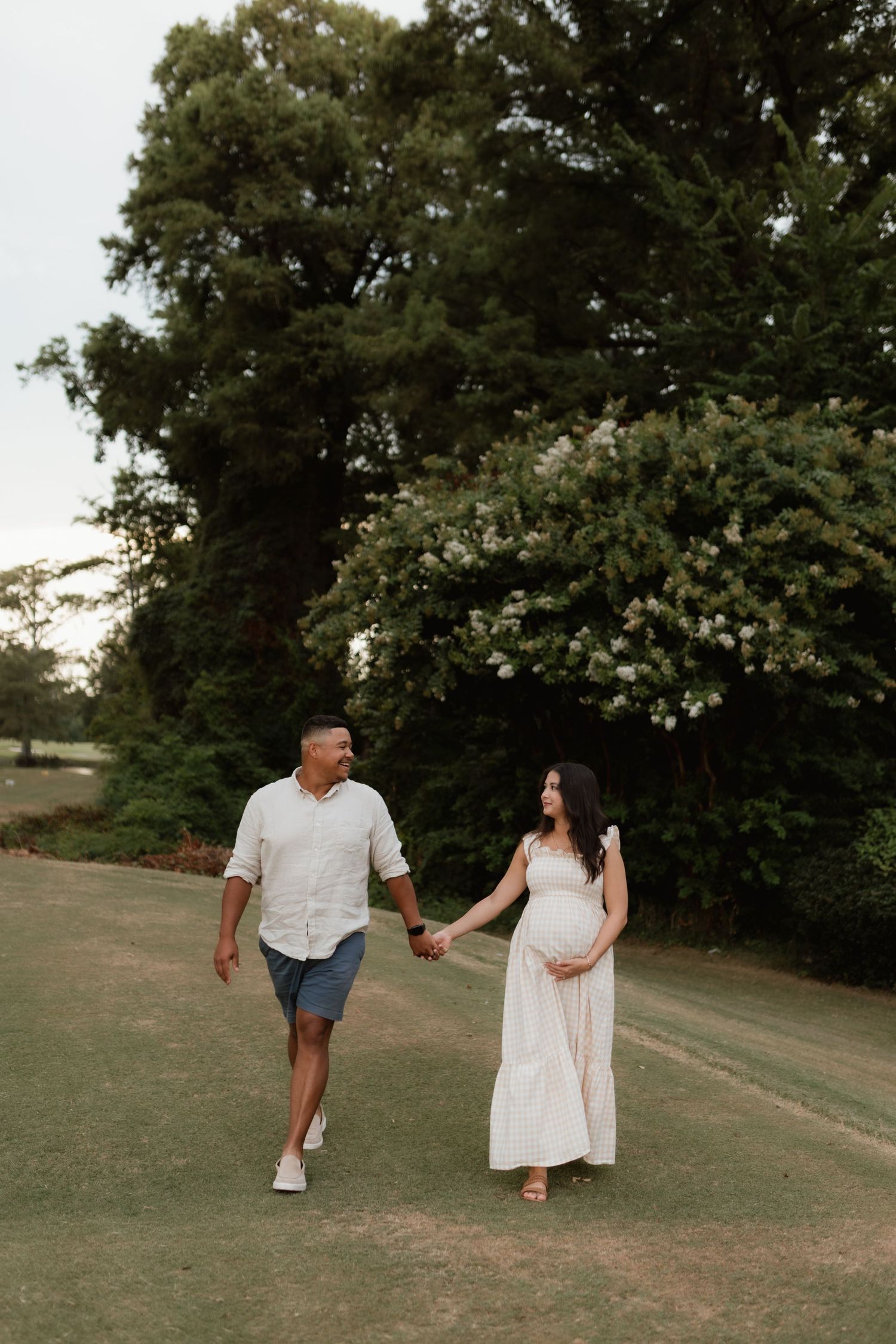 Expecting couple walking hand-in-hand on green lawn surrounded by lush trees at dusk.
