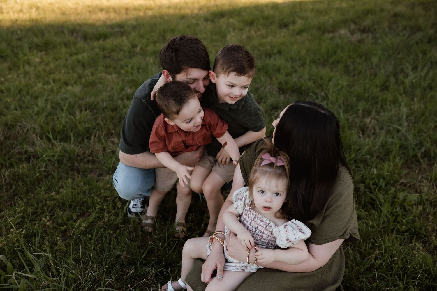 Family of five sitting on grass with parents lovingly interacting with three young children outdoors.
