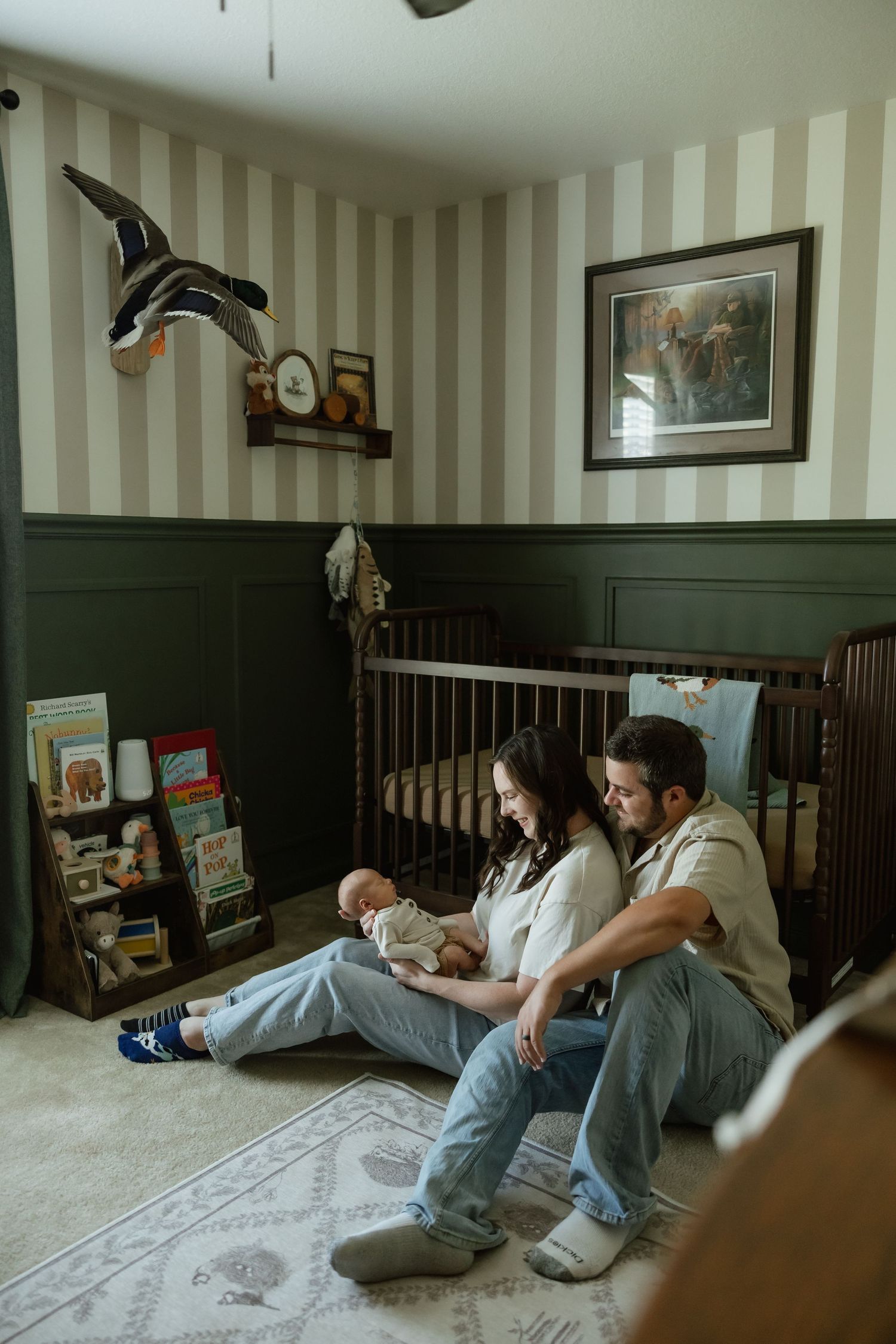 New parents sitting on nursery floor holding newborn beside a wooden crib with striped walls.