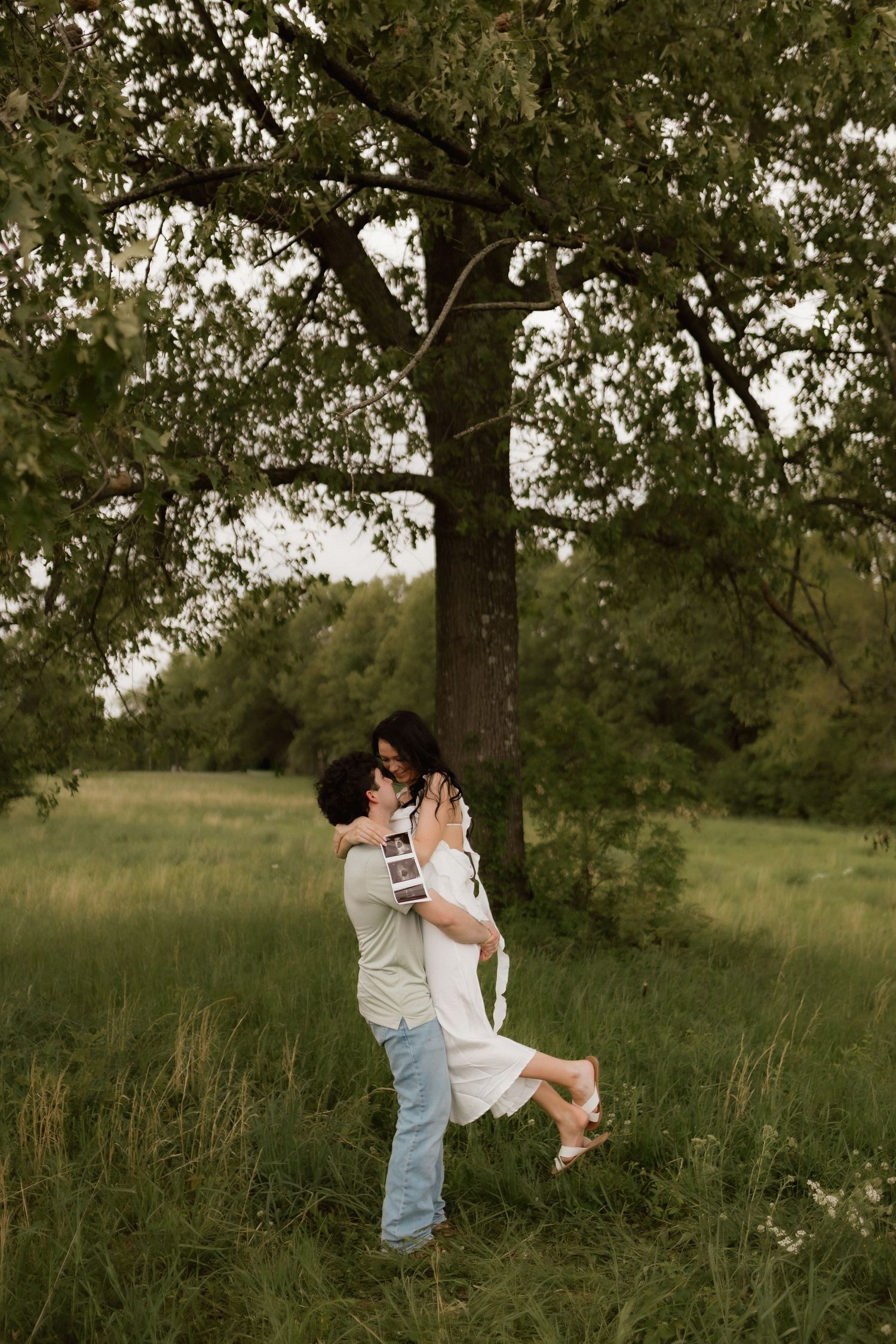 Joyful man lifting pregnant partner near tree in green field, both smiling during outdoor maternity photo session.
