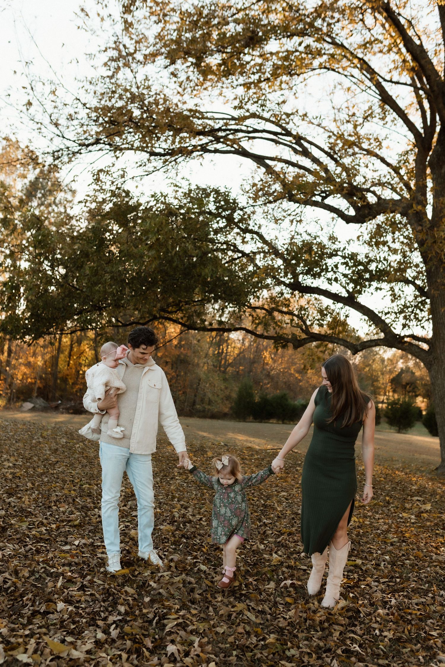 Family of four holding hands among autumn leaves under a large oak tree during golden hour in a park.