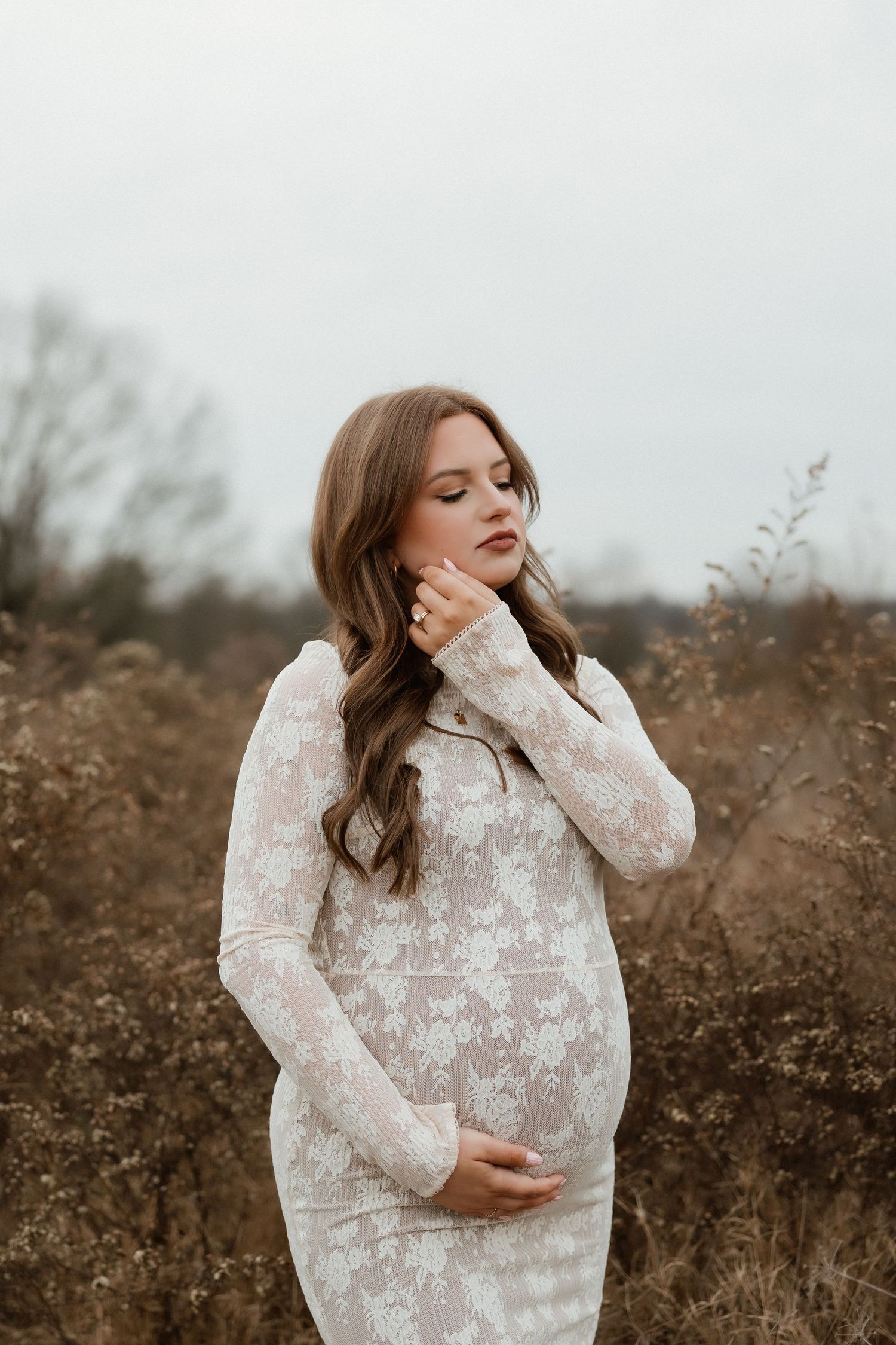 Pregnant woman in white floral lace dress cradling baby bump in a dry autumn field, eyes closed peacefully.