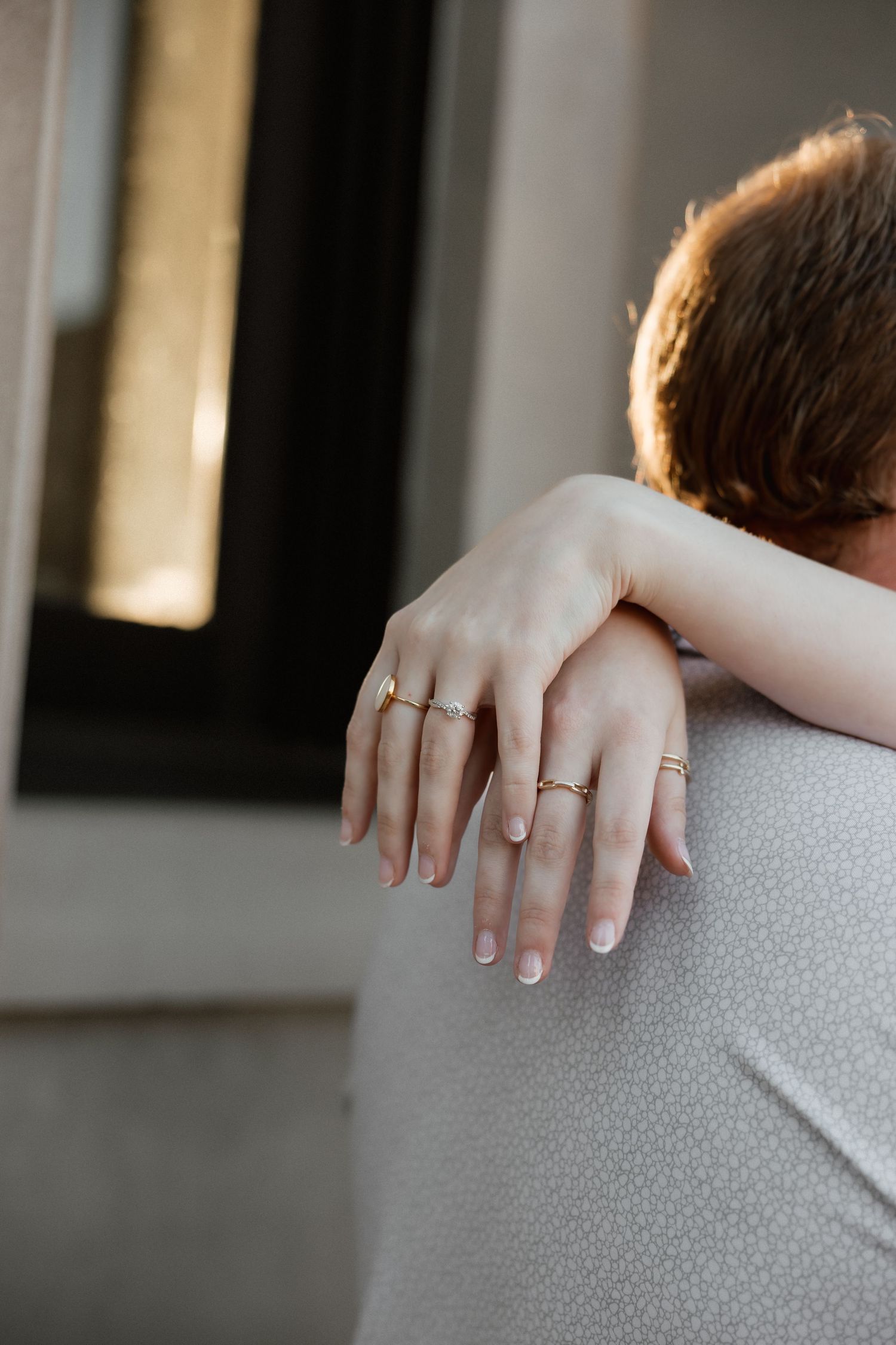 Close-up of a woman's hands adorned with gold rings wrapped around her partner's shoulder.