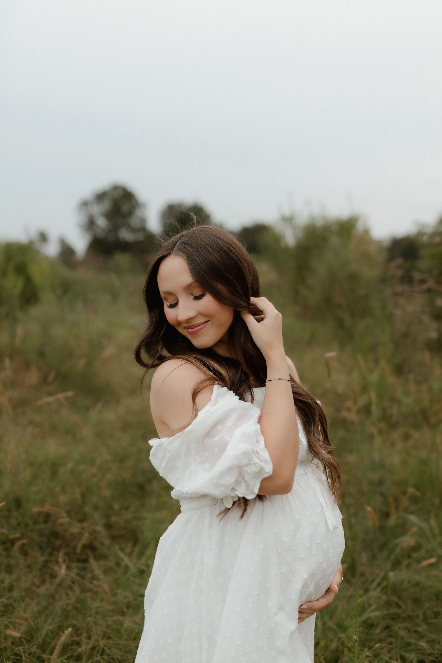 Smiling pregnant brunette in white dress gently holds her baby bump in an open meadow.